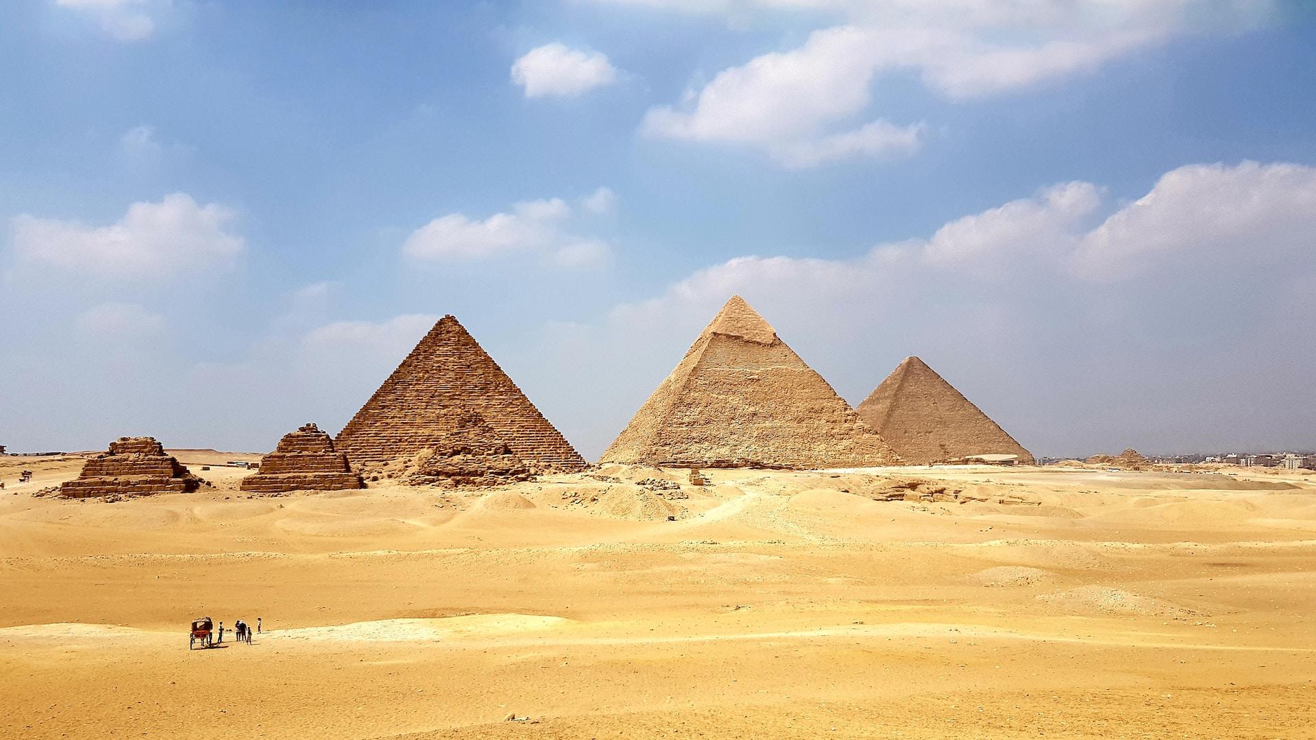 Drei große und drei kleine Pyramiden stehen auf sandigem Boden und der Himmel ist blau.