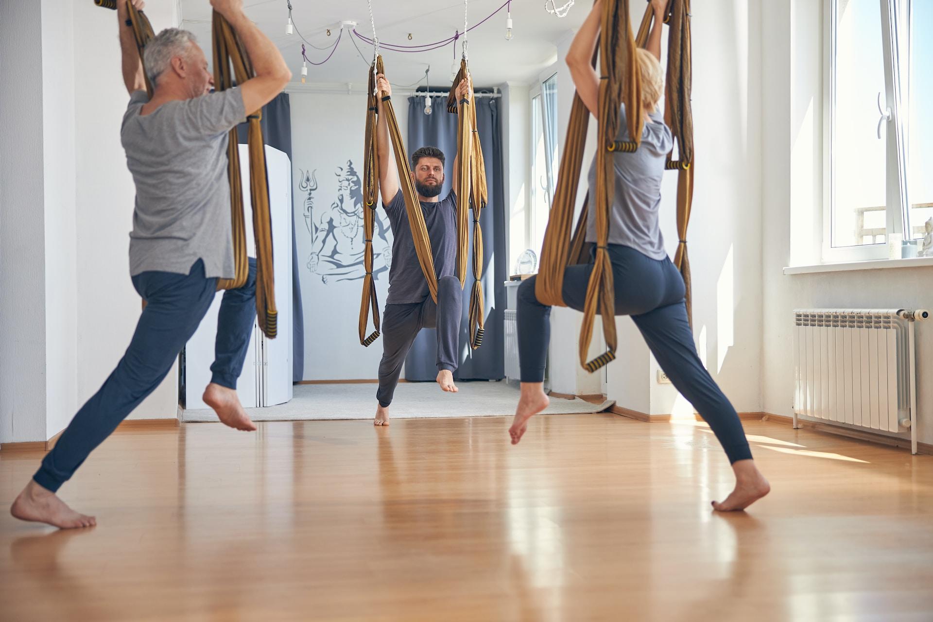 Drei Personen stehen mit einem Bein auf dem Boden und haben das andere in ein Yogatuch gehängt.