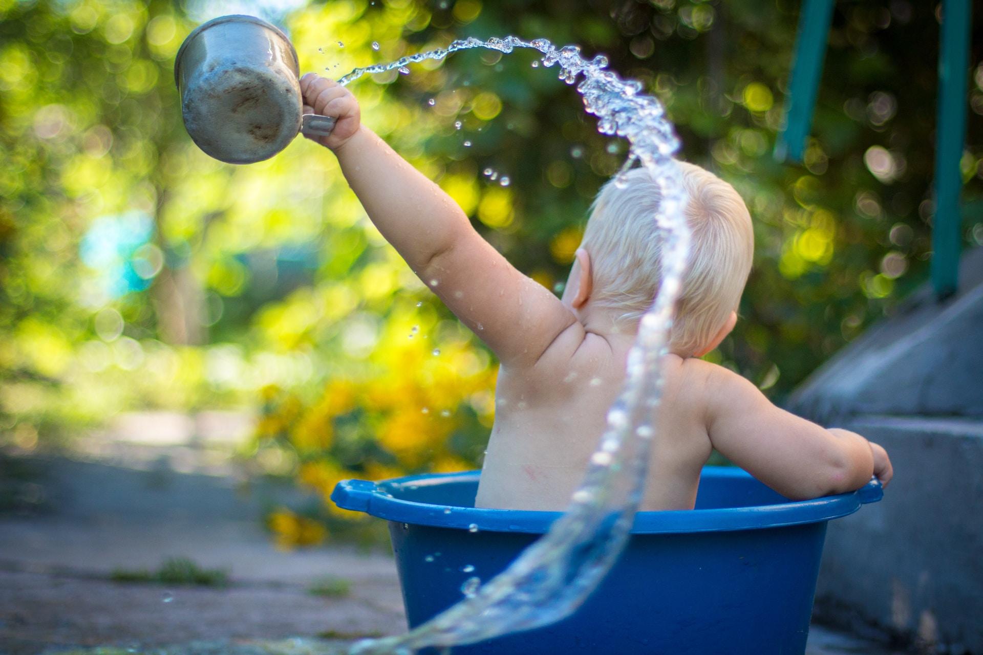 Ein Baby sitzt in einem Eimer und leert Wasser aus einem Gefäß in seiner Hand.