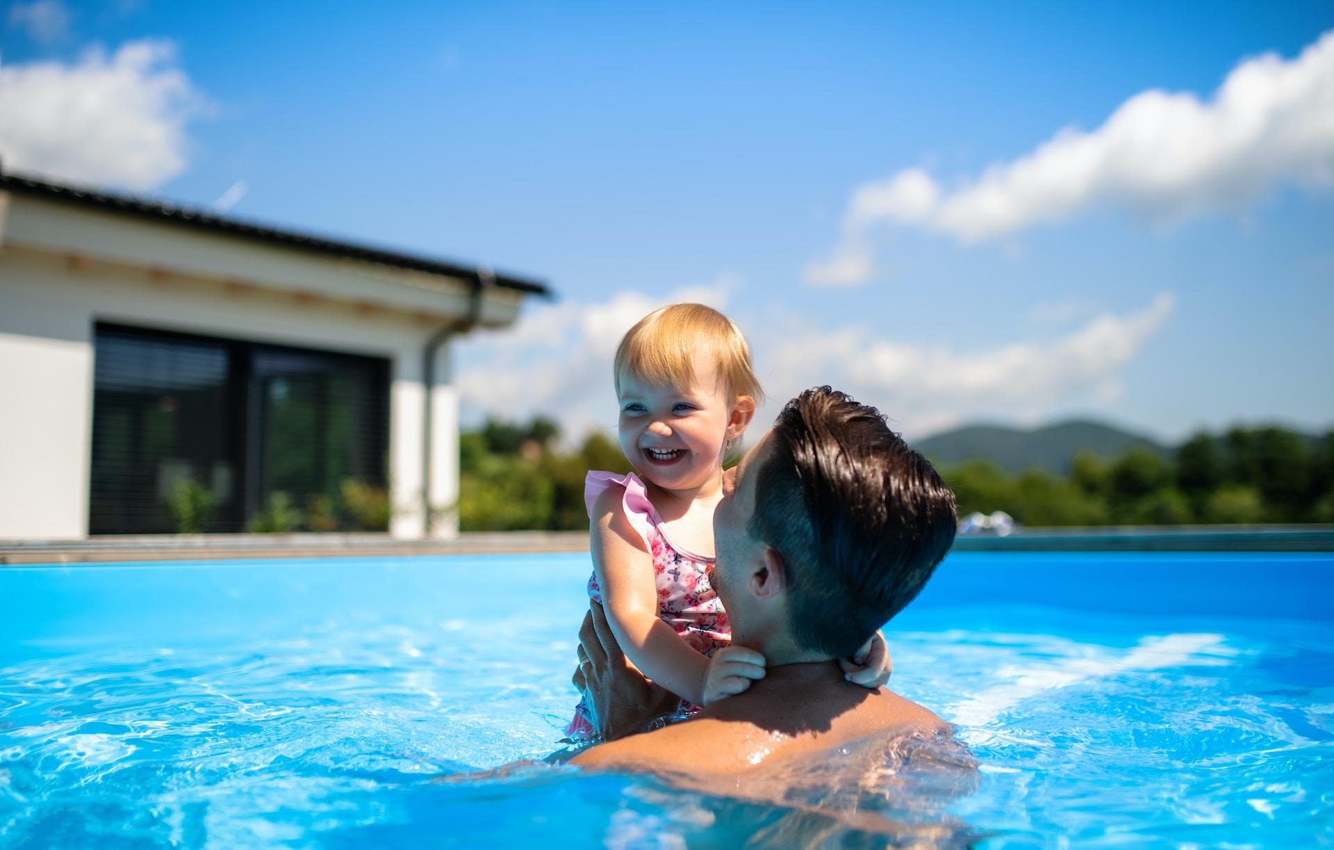 Ein Mann hält in einem Swimming Pool ein lachendes Kleinkind in die Höhe.