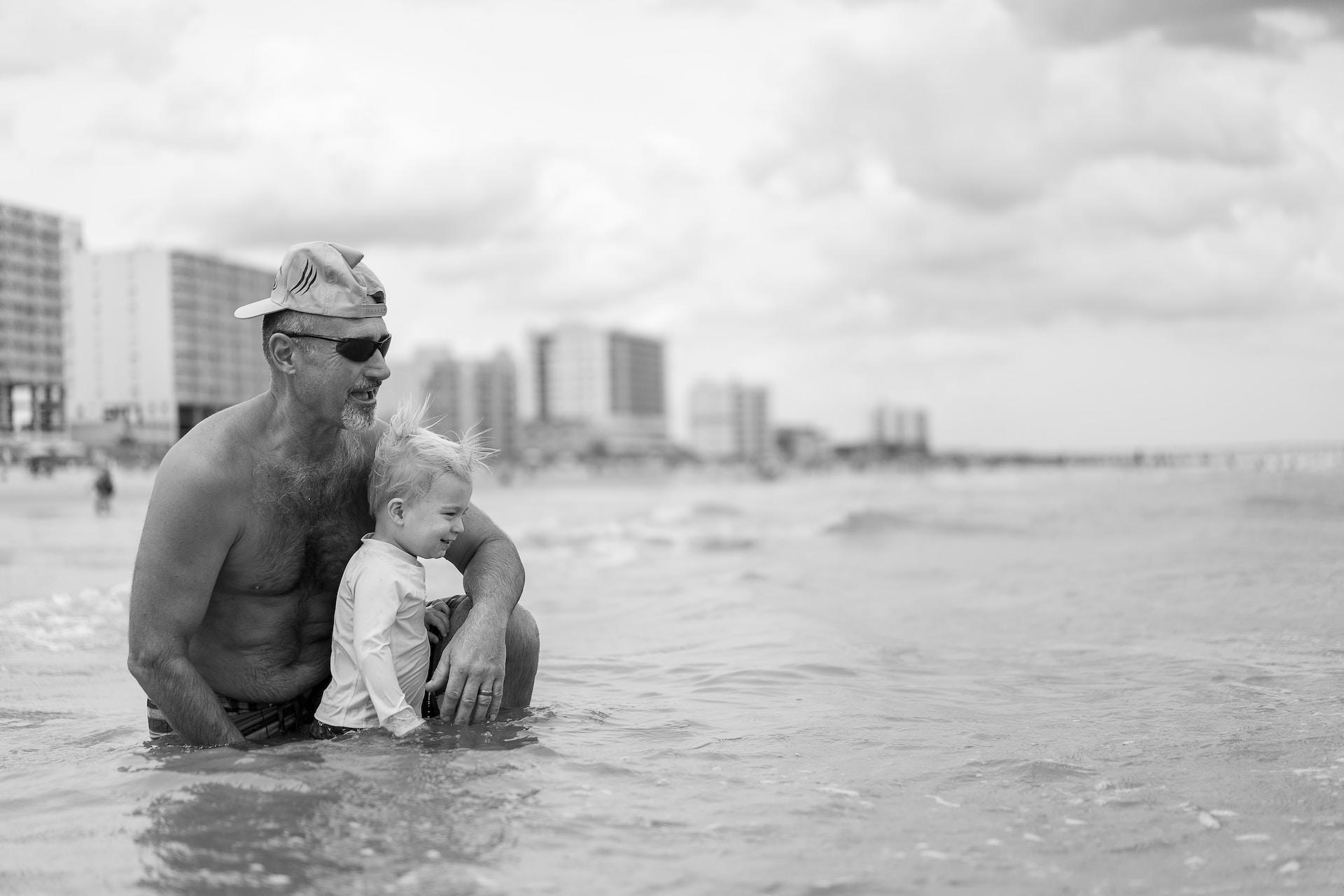 Eine erwachsene Person sitzt mit einem Kleinkind im Meer vor großen Häusern.