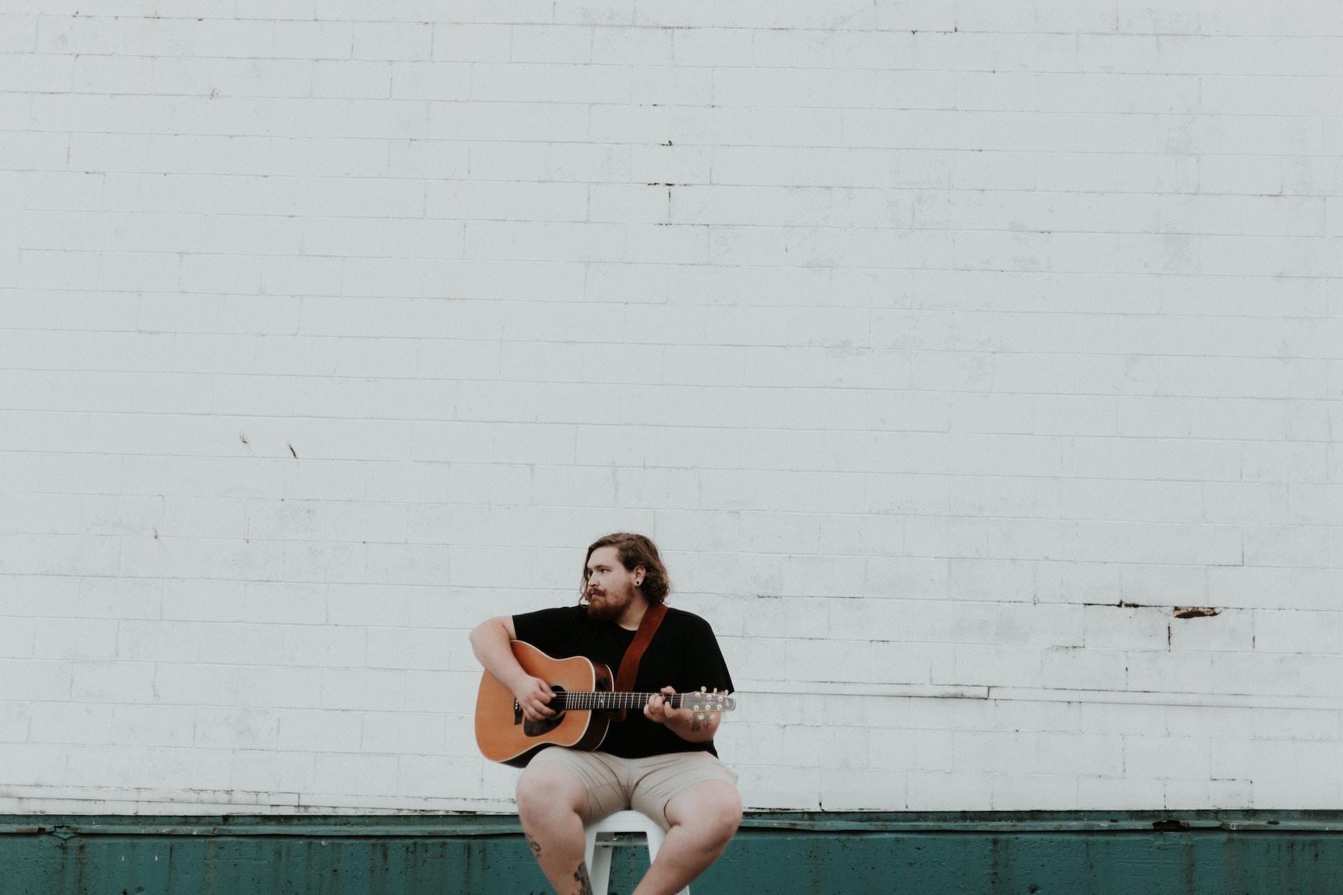 Ein Sänger gibt sitzend auf einem Hocker vor einer weißen Wand mit Gitarre ein Lied zum besten.