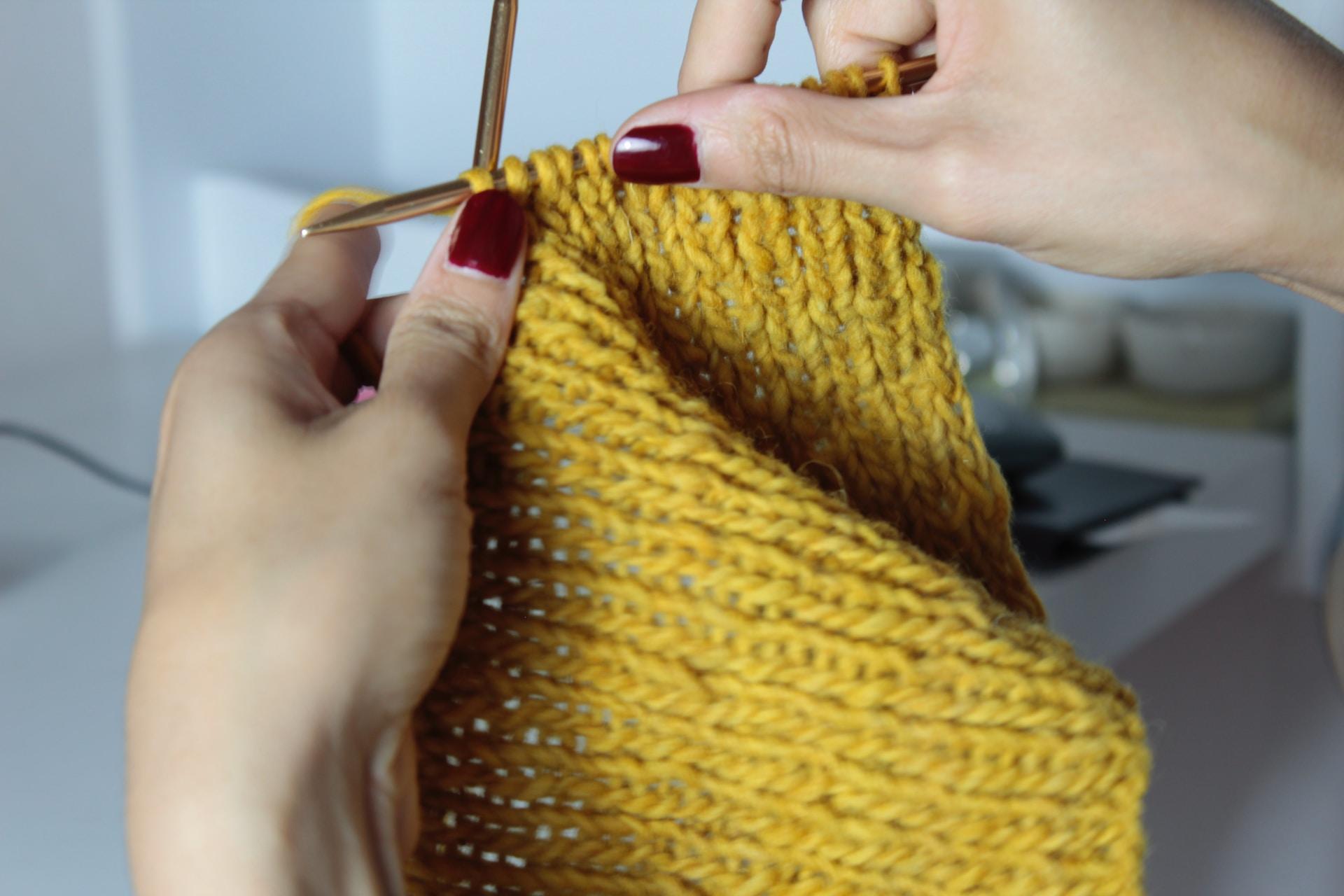 Zwei Hände halten zwei Stricknadeln und gelben Garn fest.
