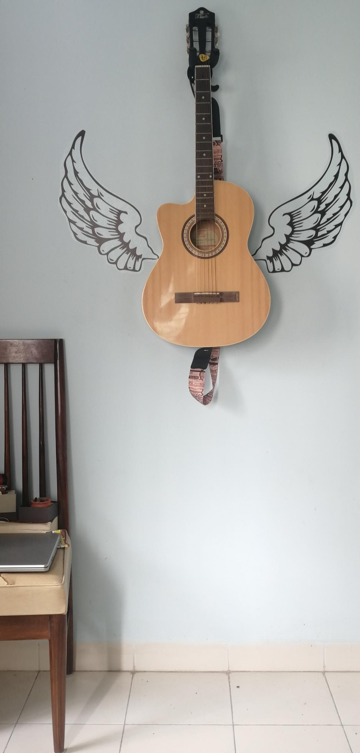 Eine Gitarre hängt an einer Wand mit aufgemalten Engelsflügeln und daneben steht ein Stuhl