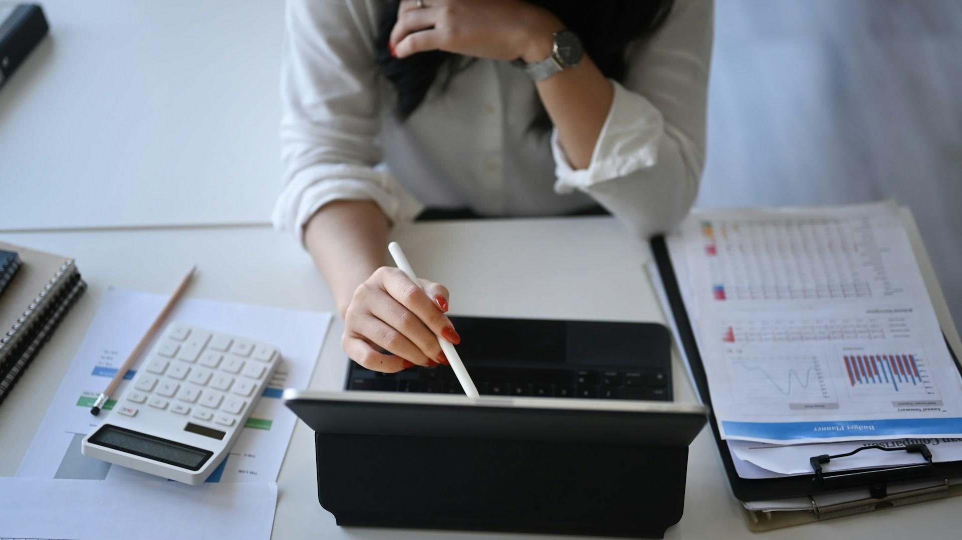 Eine Person am Laptop und daneben liegen Listen und ein Taschenrechner