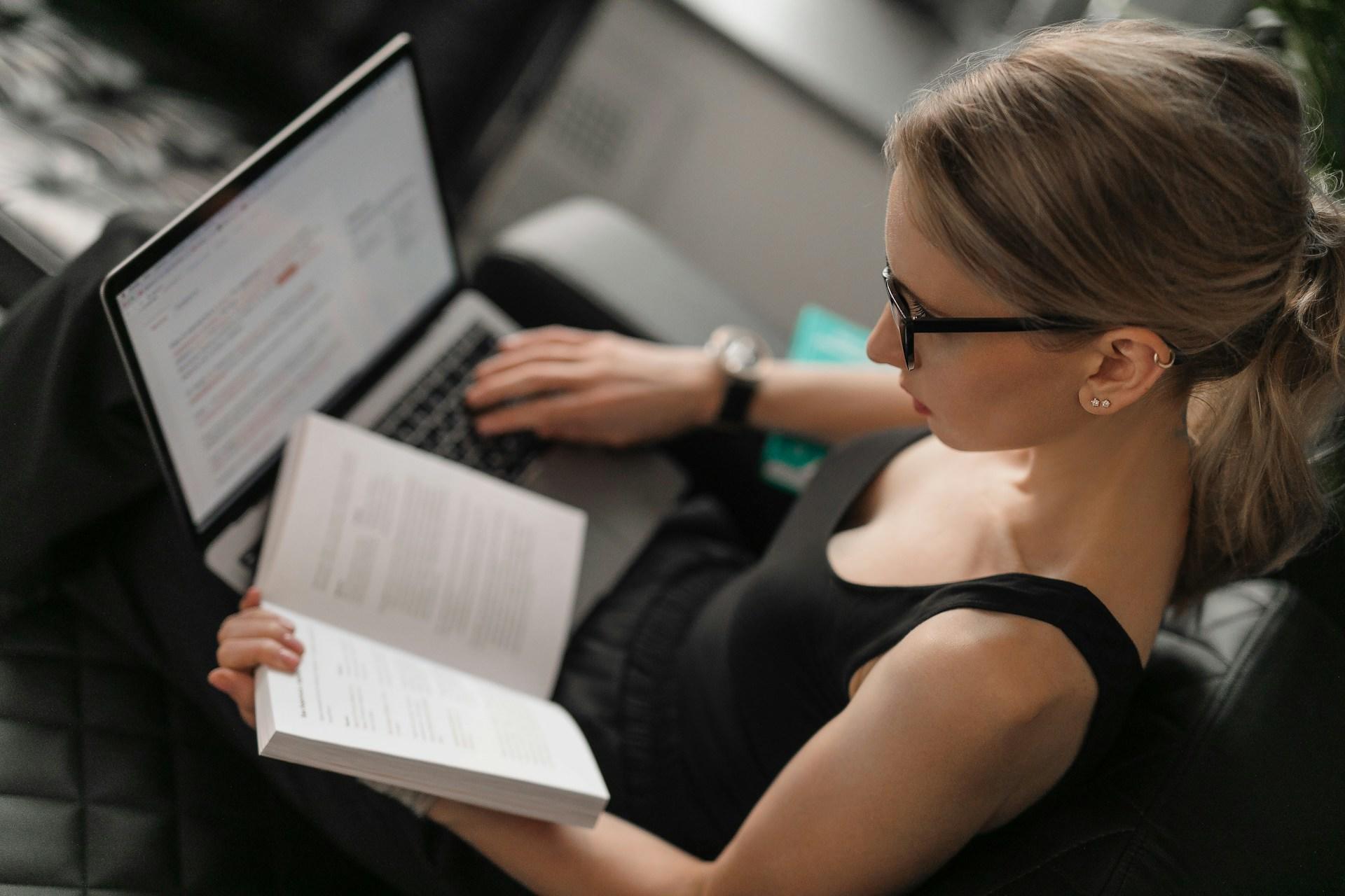 Frau mit Buch in der Hand und Laptop auf dem Schoß