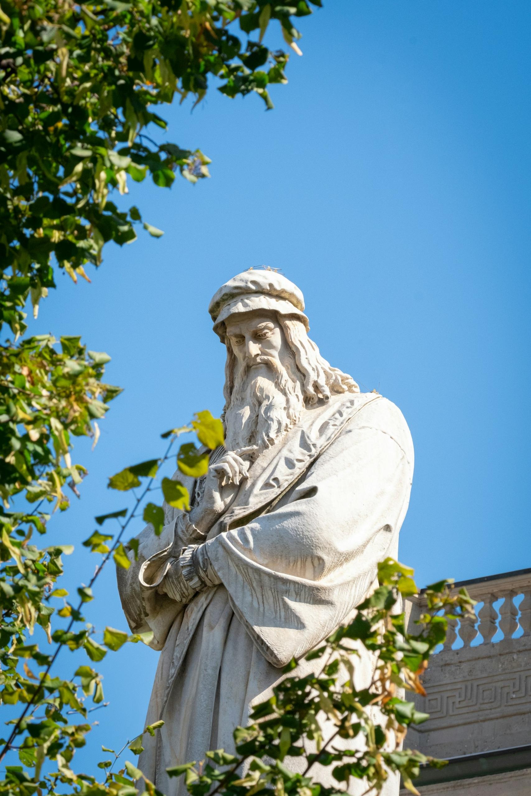 Statue von Mann mit Mütze vor blauem Himmel und Baum.