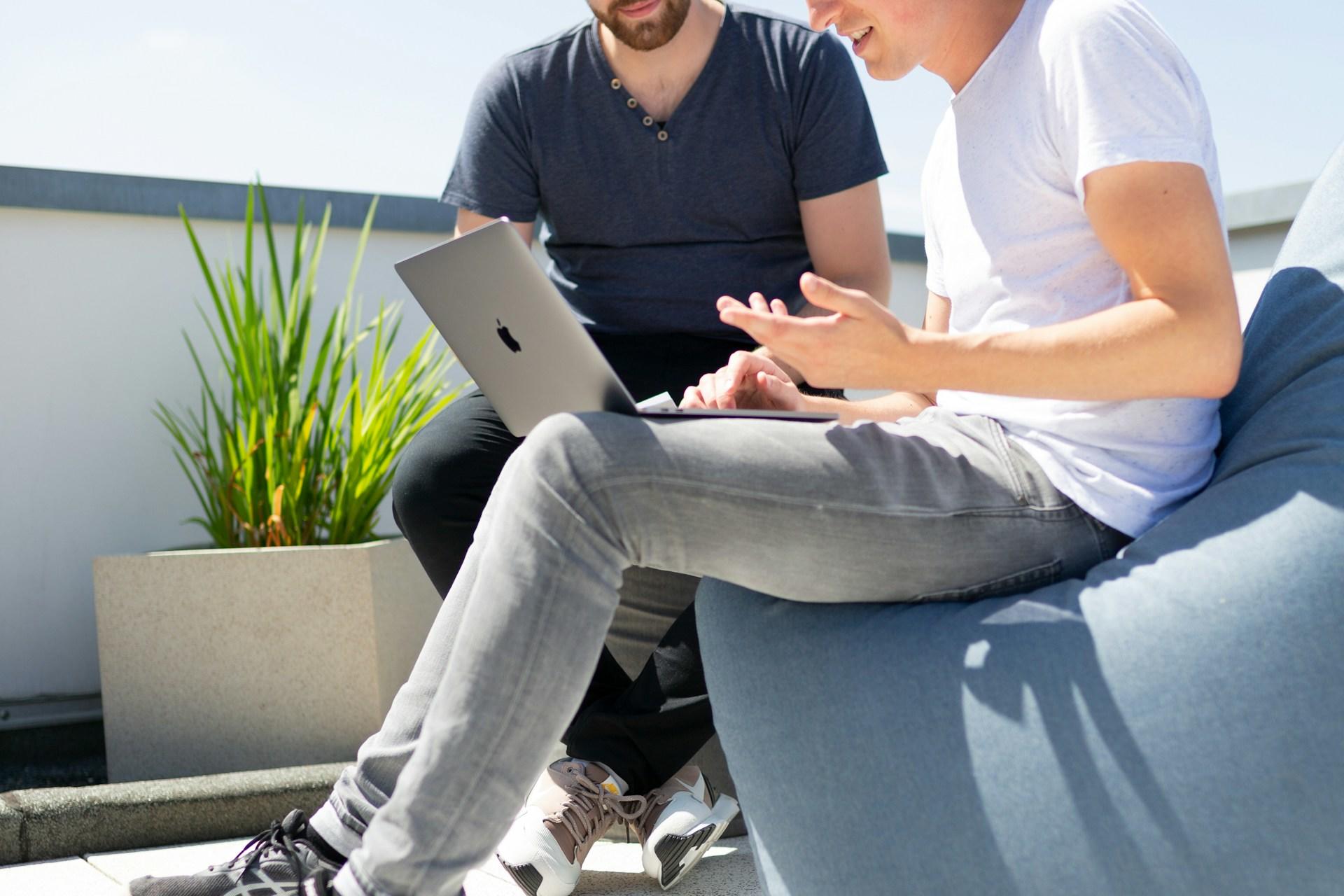 Zwei Männer sitzen nebeneinander auf Sitzkissen mit Laptop in der Hand.