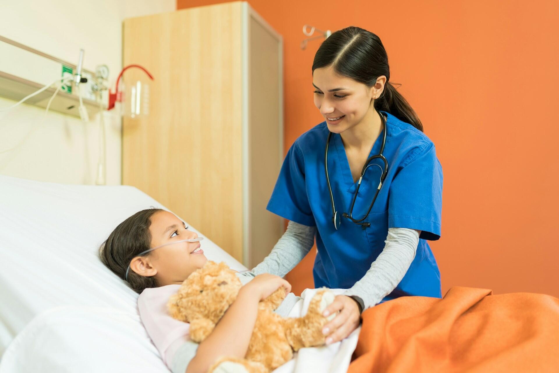 Ein Kind liegt mit Teddy im Arm in einem Krankenhausbett und daneben steht eine erwachsene Person