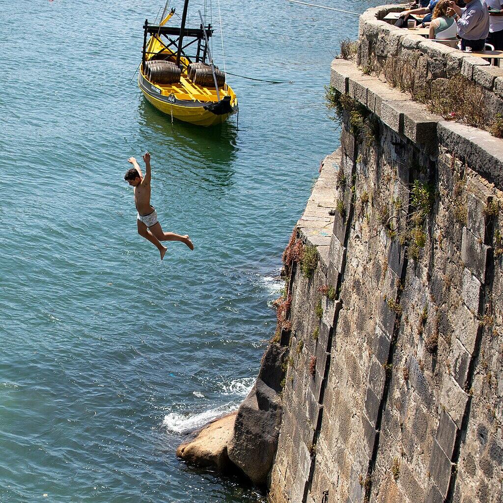 Person springt von einer hohen Mauer ins Meer und dahinter ist ein gelbes Boot