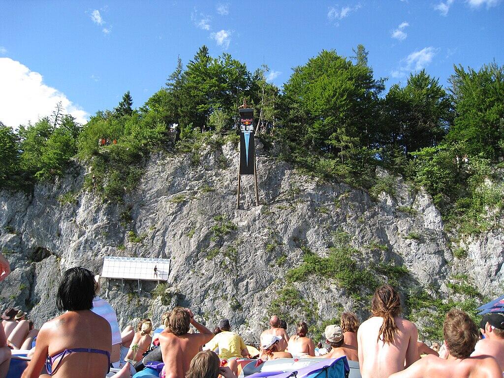 Einige Zuschauer sehen zu einer Klippe auf. Ein Springer steht dort auf einem absprungbrett mit dem Red Bull Cliff Diving Logo.