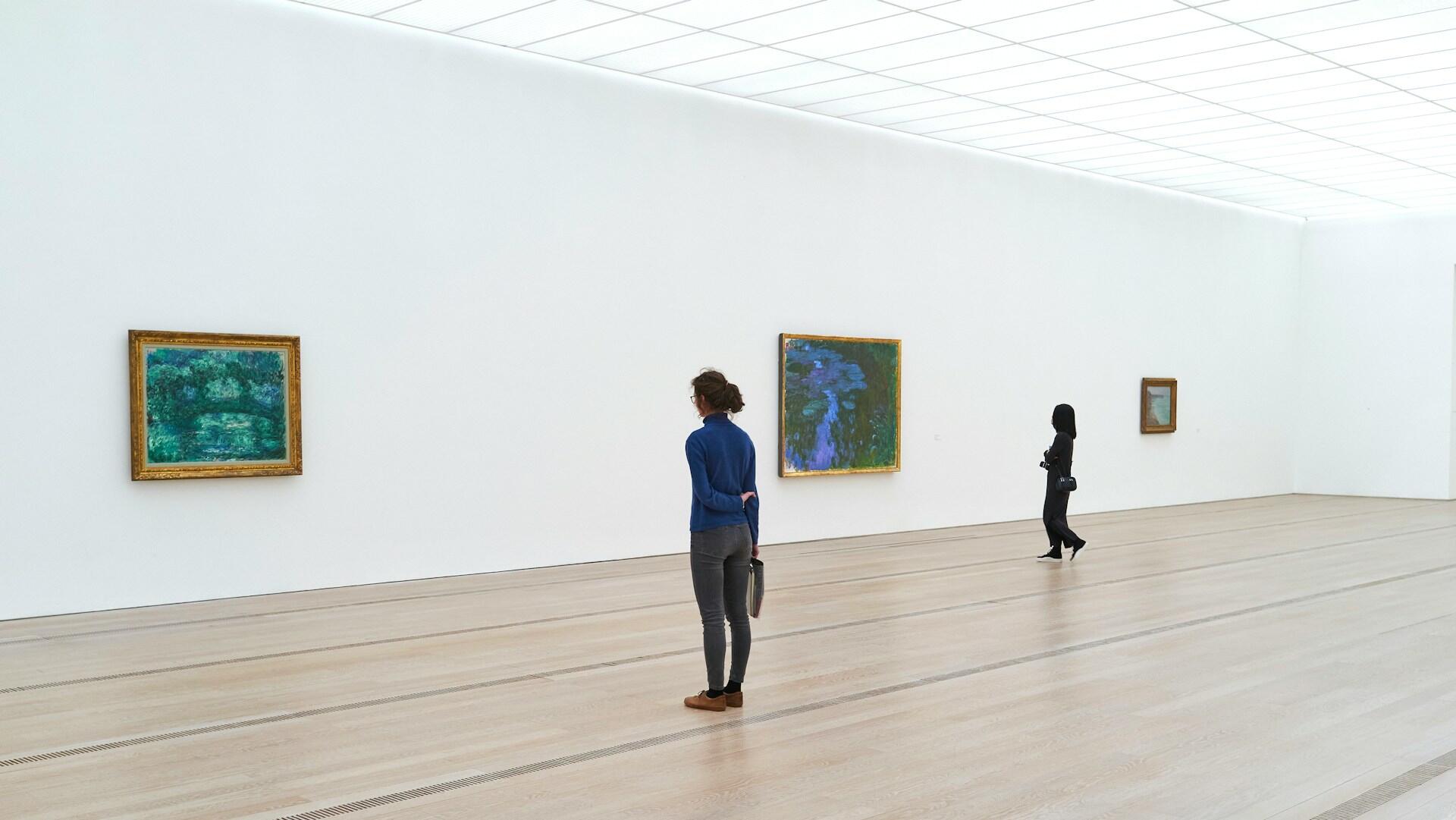 Zwei Menschen stehen in einer Galerie mit großen weißen Wänden, in der 3 Bilder ausgestelt sind.