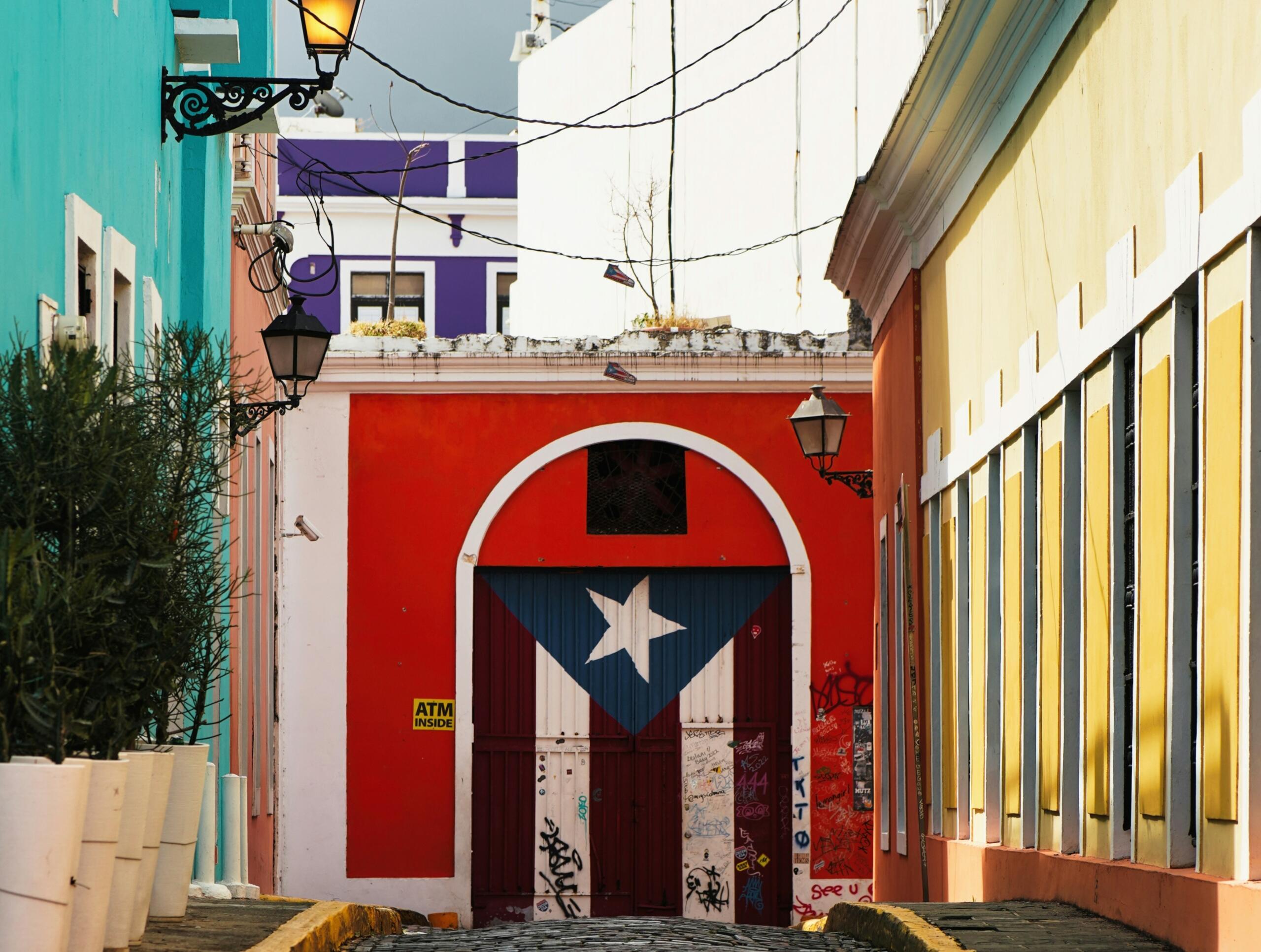 Eine lebendige Gasse mit bunten Häusern, featuring eine rote Tür, geschmückt mit der Flagge von Puerto Rico, und markante gelbe Fensterläden.