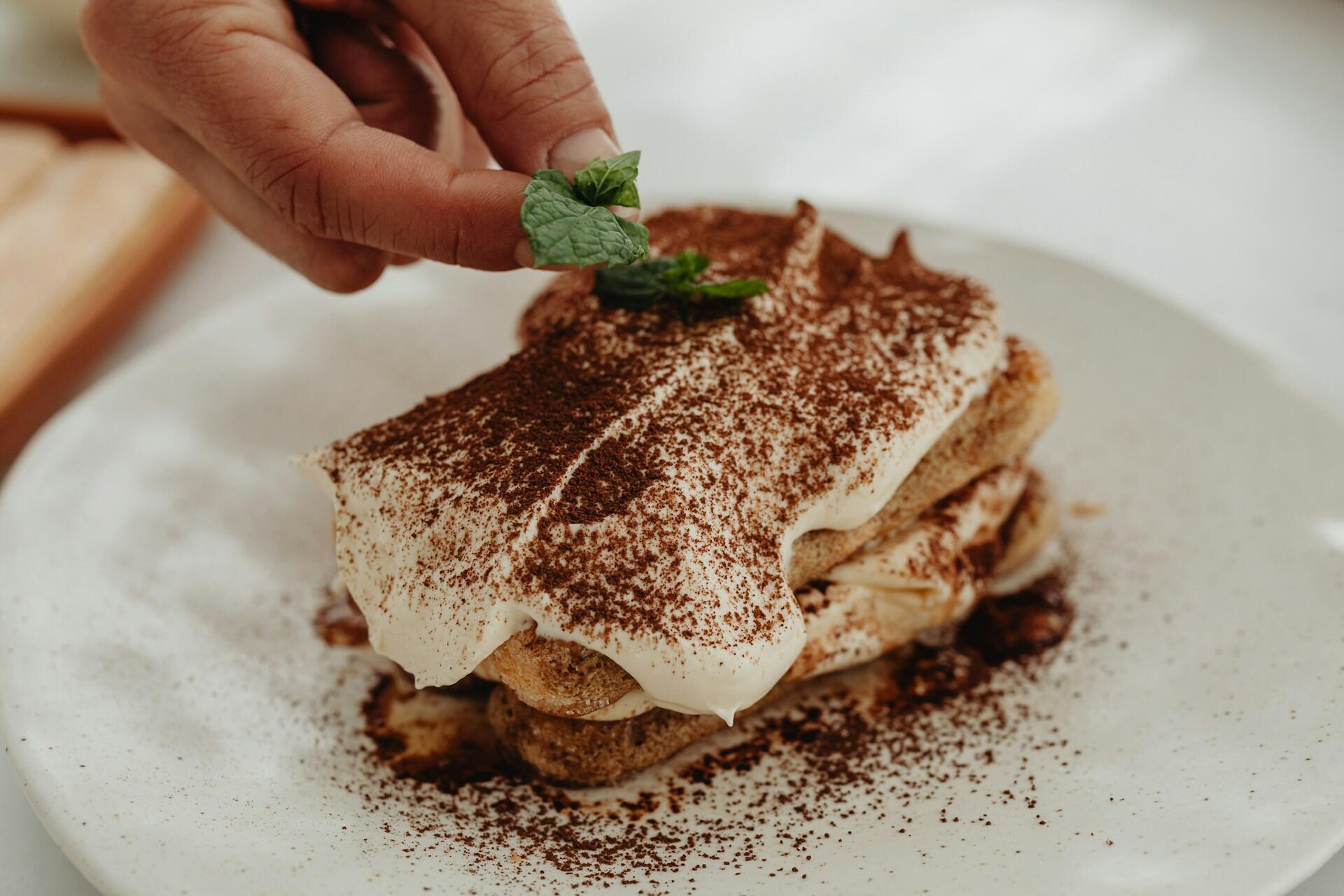 Eine Hand dekoriert ein köstliches Tiramisu-Dessert in Schichten mit Kakaopulver und frischen Minzblättern auf einem weißen Teller.