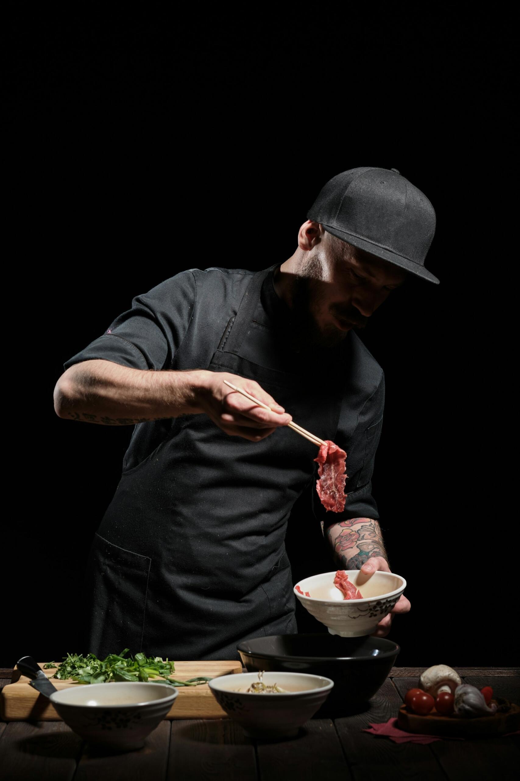 Ein Koch mit schwarzer Schürze hält rohes Fleisch mit Stäbchen über eine Schüssel, umgeben von frischen Zutaten auf einem Holztisch.