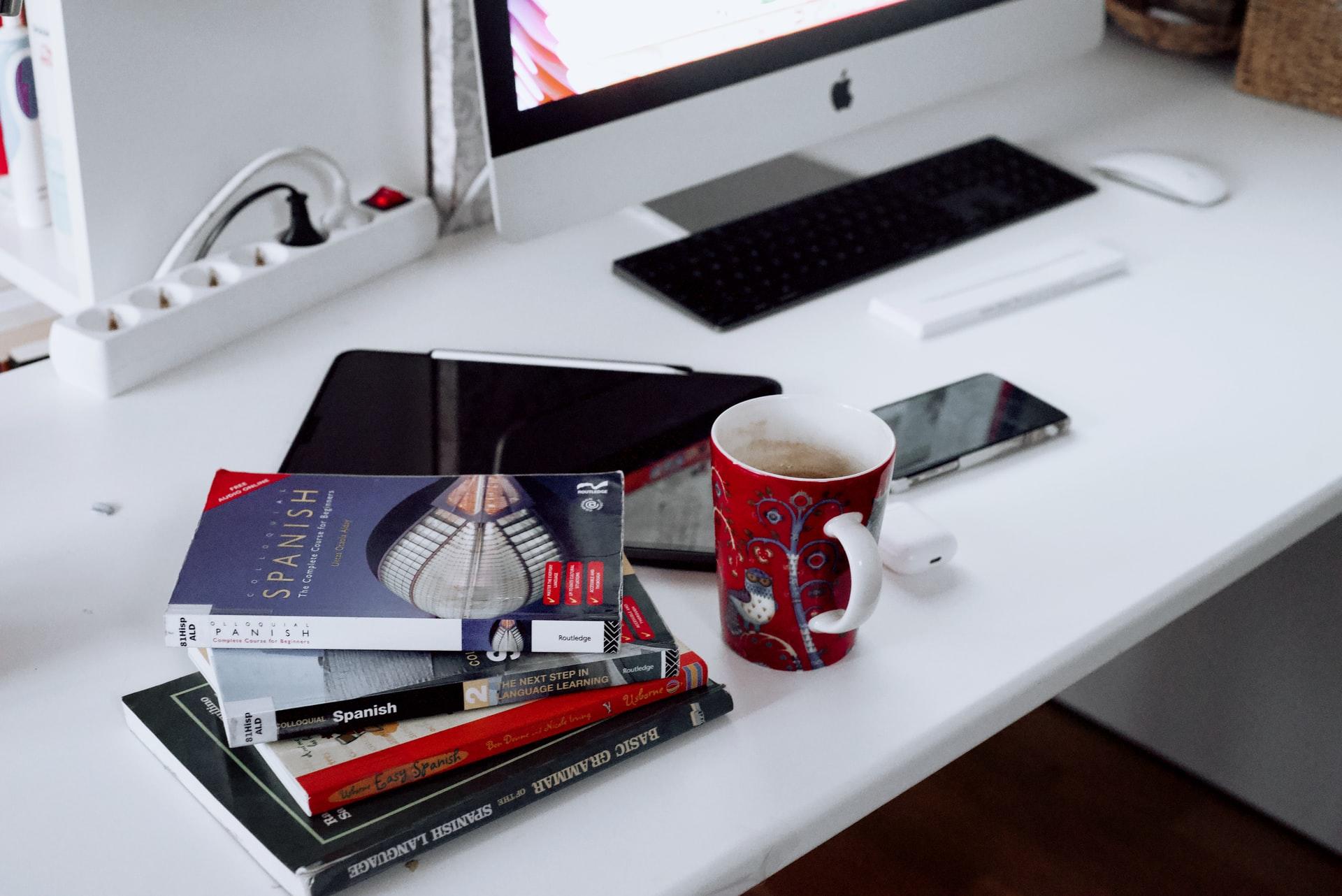Et skrivebord med fire bøger, en iPad, AirPods, en iPhone, en iMac og en kaffekop