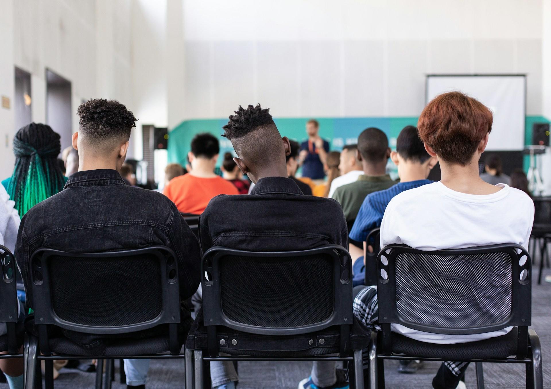 Junge Menschen sitzen in Reihen auf Stühlen und blicken auf Lehrer vorne.