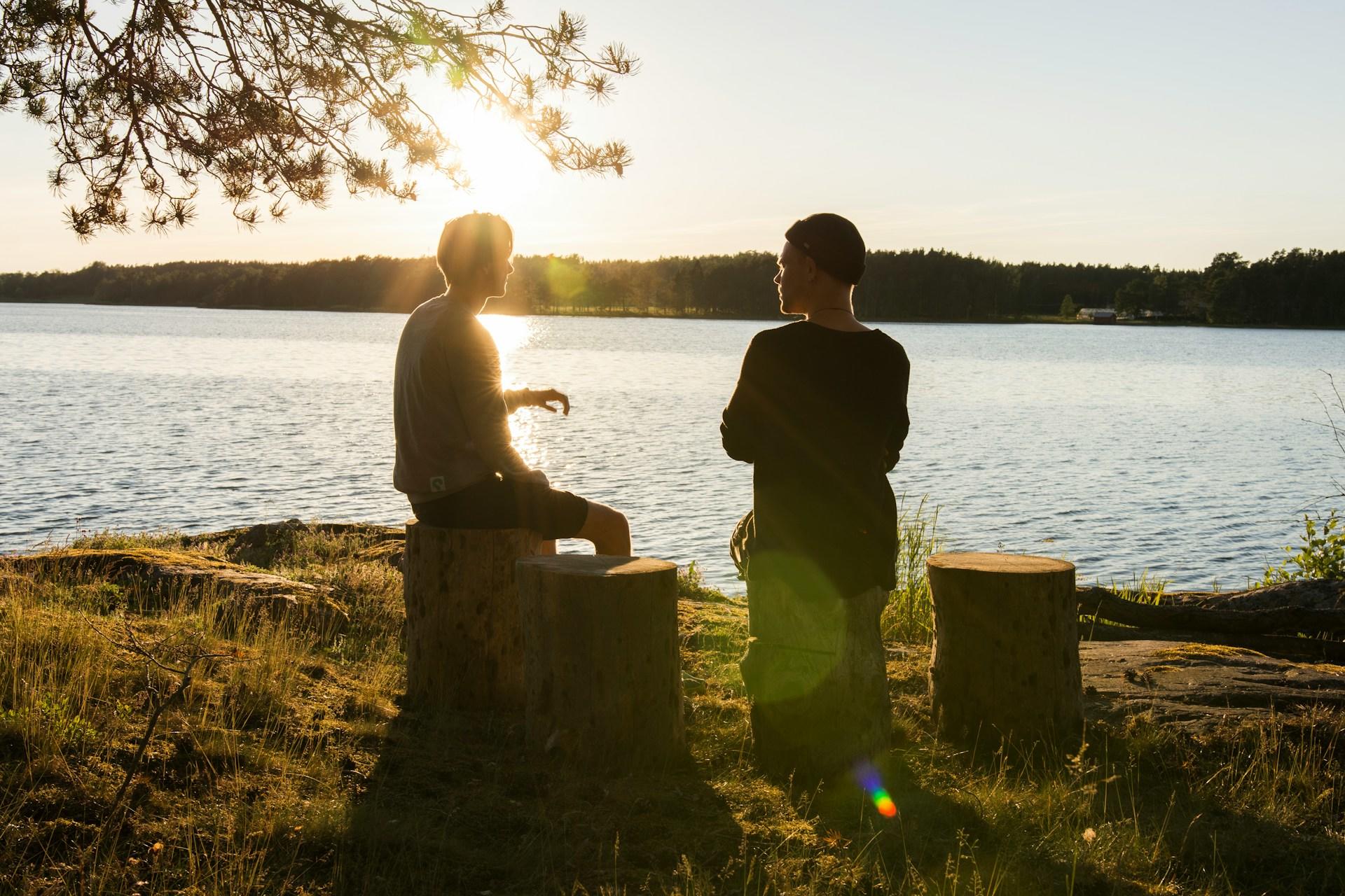 Zwei Männer sitzen auf Holzstämmen an einem See.