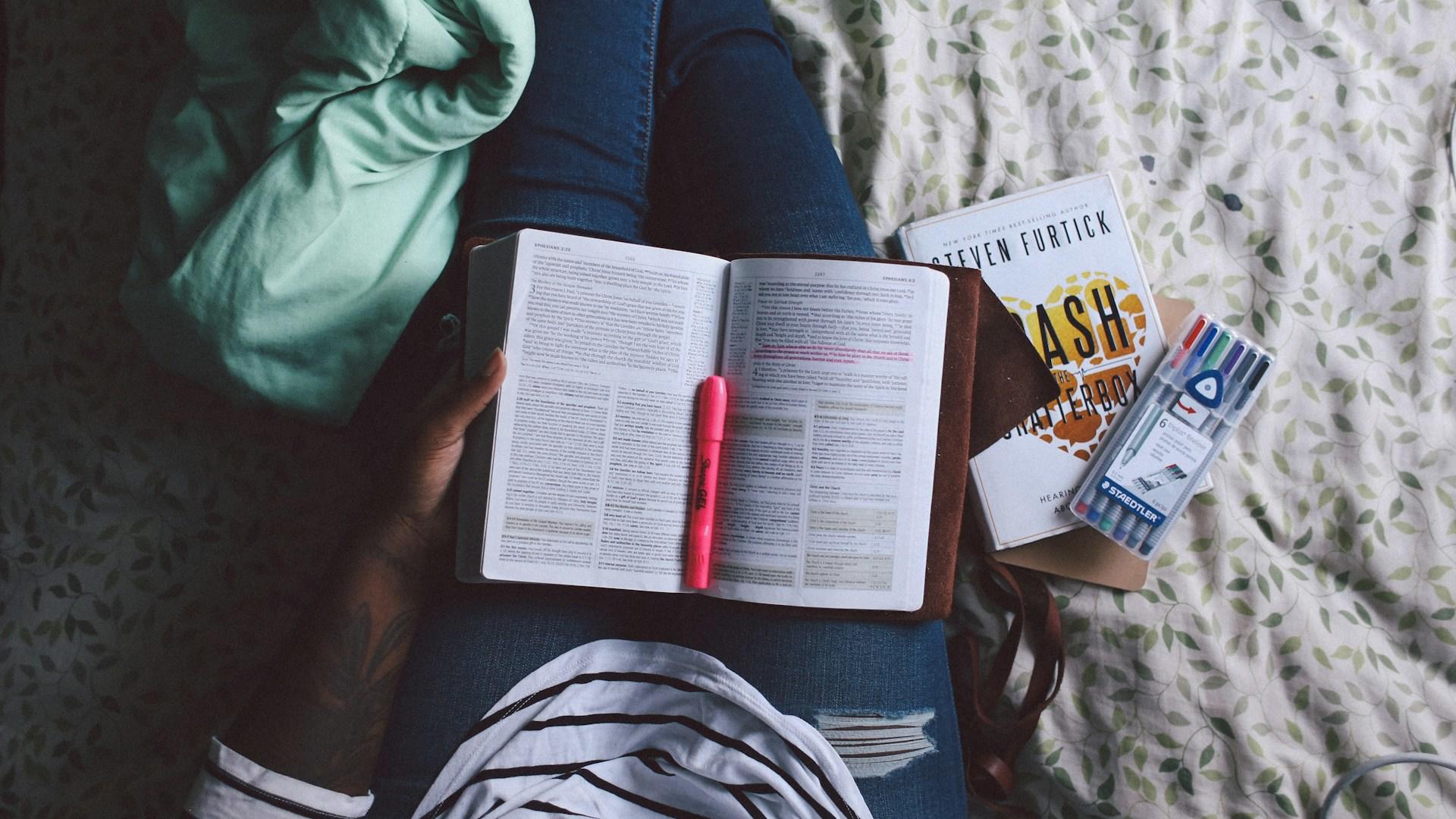 Person sitzt auf Bett mit aufgeschlagenem Buch und Textmarker.
