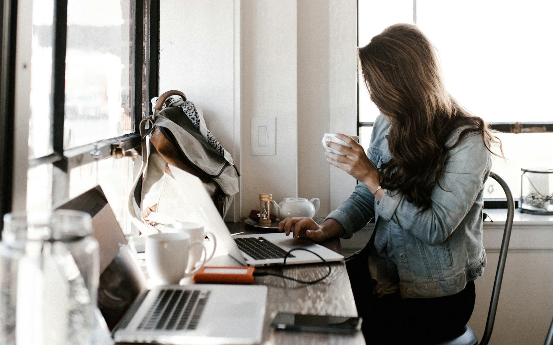 Eine junge Frau sitzt vor ihrem Laptop und trinkt Kaffee