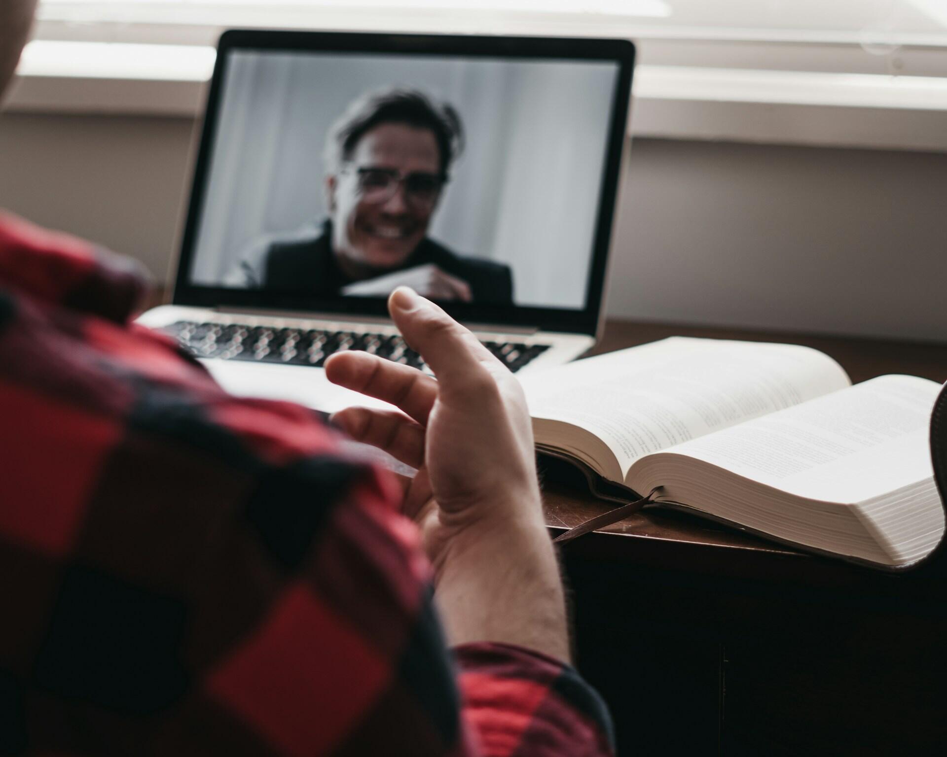 Ein junger Mann sitzt vor einem Laptop in einem online Meeting, wo ein anderer Mann zu sehen ist. Daneben liegt ein aufgeschlagenes Buch.