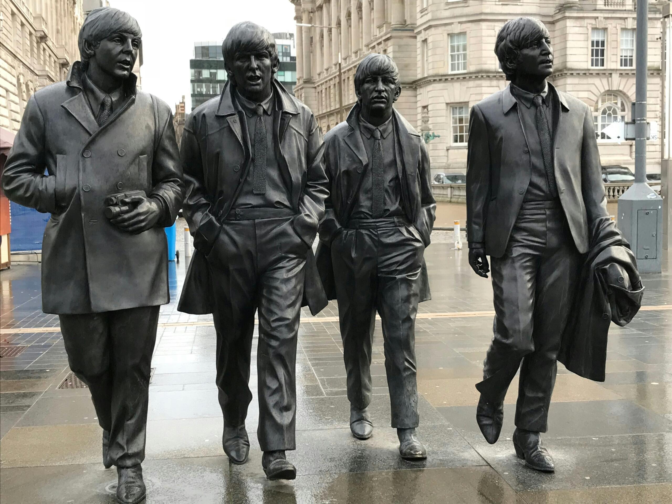 The Beatlesi liikmete pronkskujud Liverpoolis linnahoonete ees vihmasel tänaval.