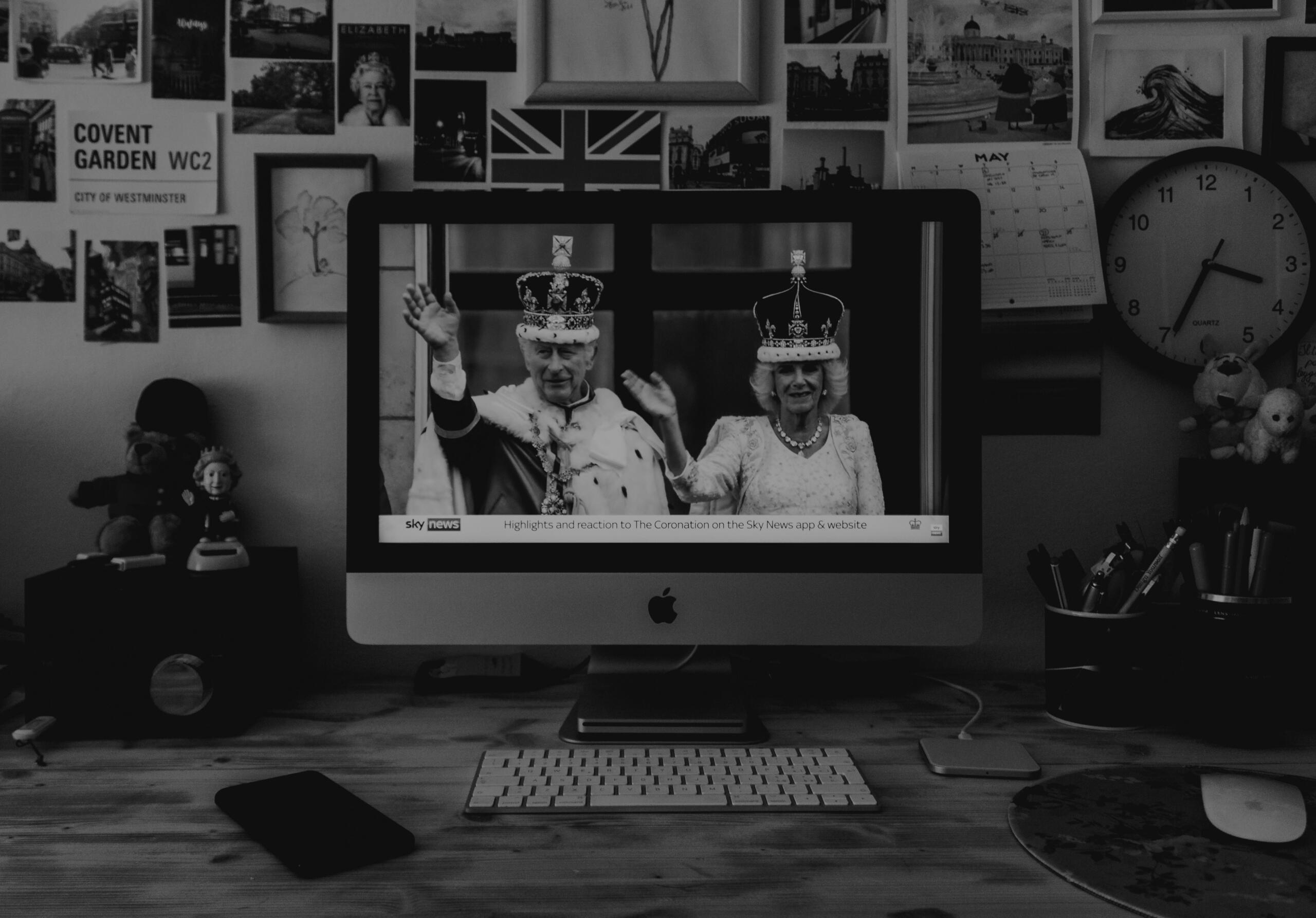 Mustvalge foto töölaust, mille arvutiekraanil on näha Briti kuninglikku kroonimistseremooniat ning taustal Ühendkuningriigiga seotud pildid ja sümbolid.