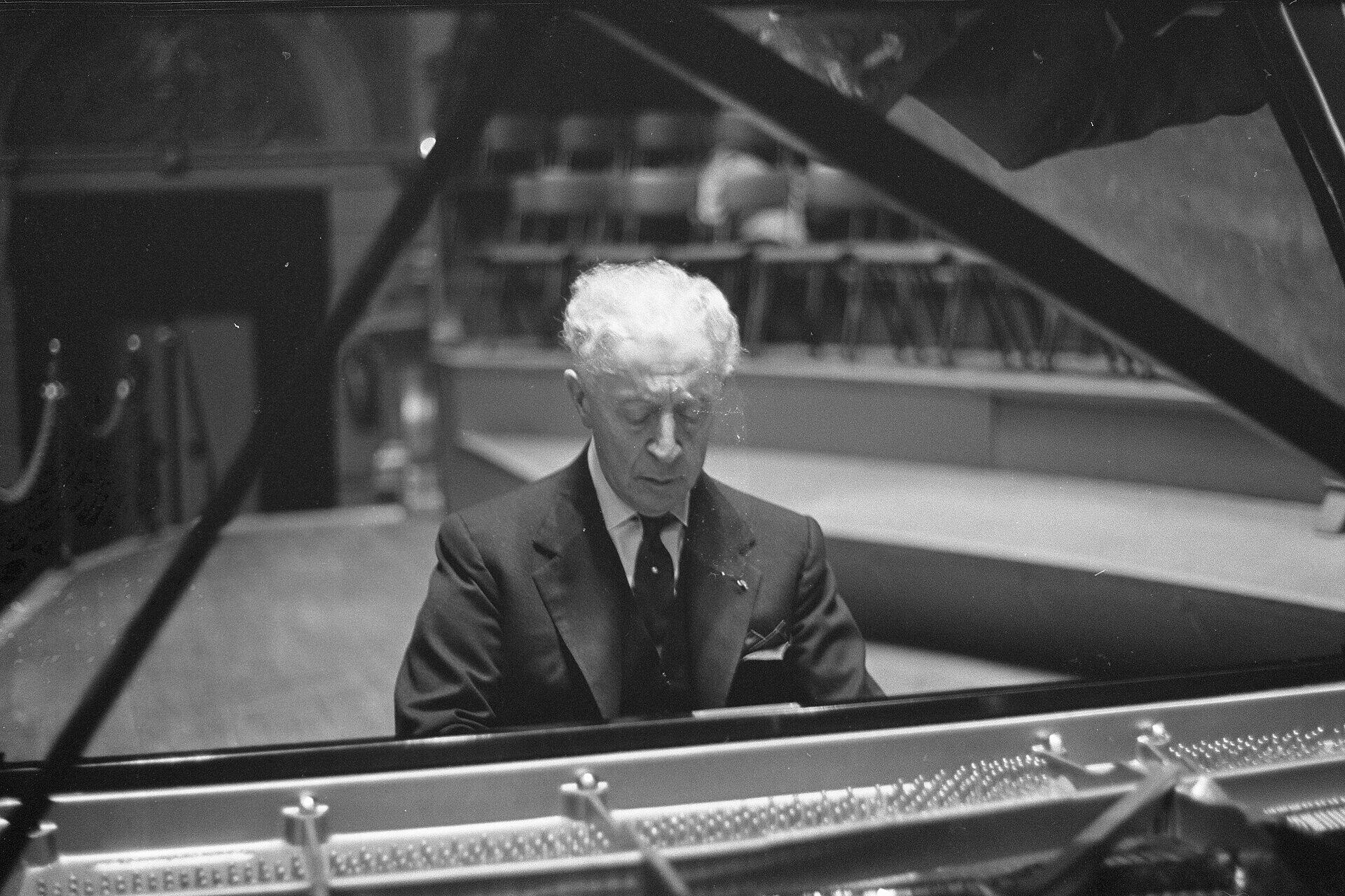 Arthur Rubinstein mängib kontserdisaalis tiibklaverit, pilk klahvidele suunatud. Foto on tehtud läbi avatud klaverikaane.