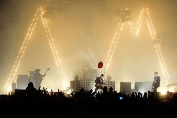 Arctic Monkeys esinemas suurel festivalilaval, taustal heledad kolmnurksed valguskonstruktsioonid ja ees publik.