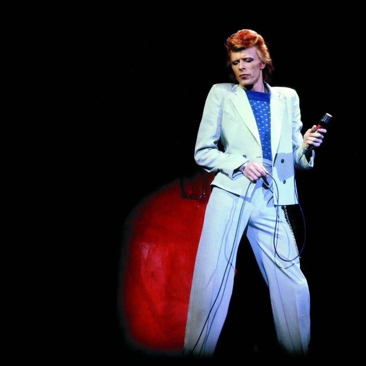David Bowie esineb laval heledas ülikonnas ja hoiab käes mikrofoni, taust on tume.