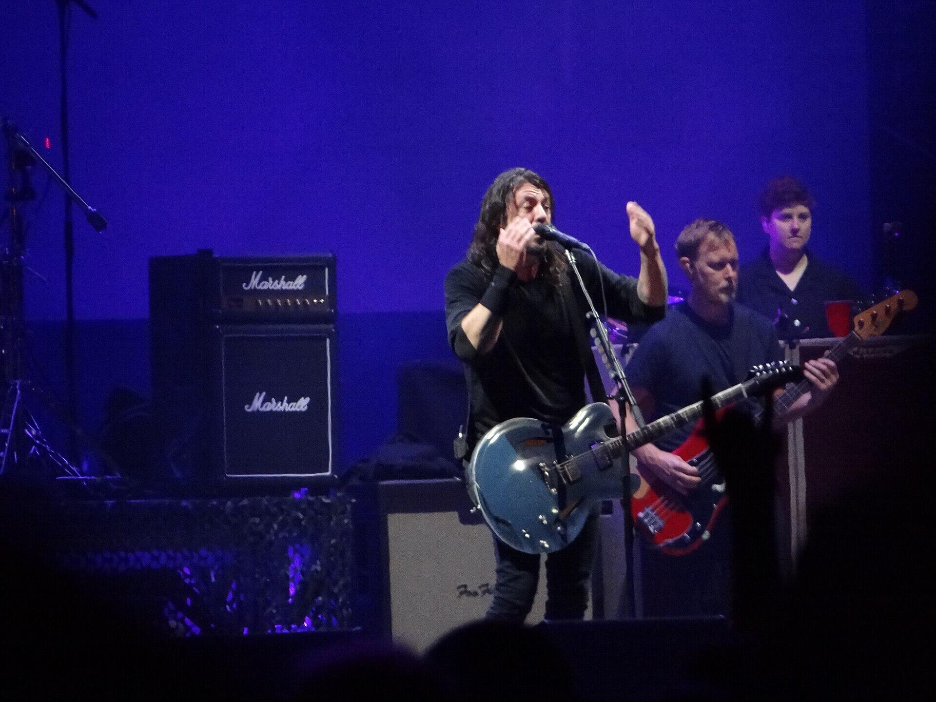 Foo Fighters esineb sinakaslilla lavavalguse all, ees laulja kitarriga ning taustal teised bändiliikmed ja võimendid.