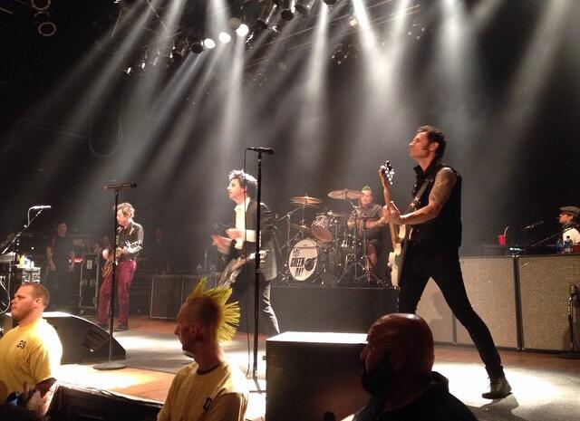 Green Day esineb laval ereda prožektorvalguse all, bändiliikmed mängivad pille ning lava ees on näha publikut ja turvatöötajaid.