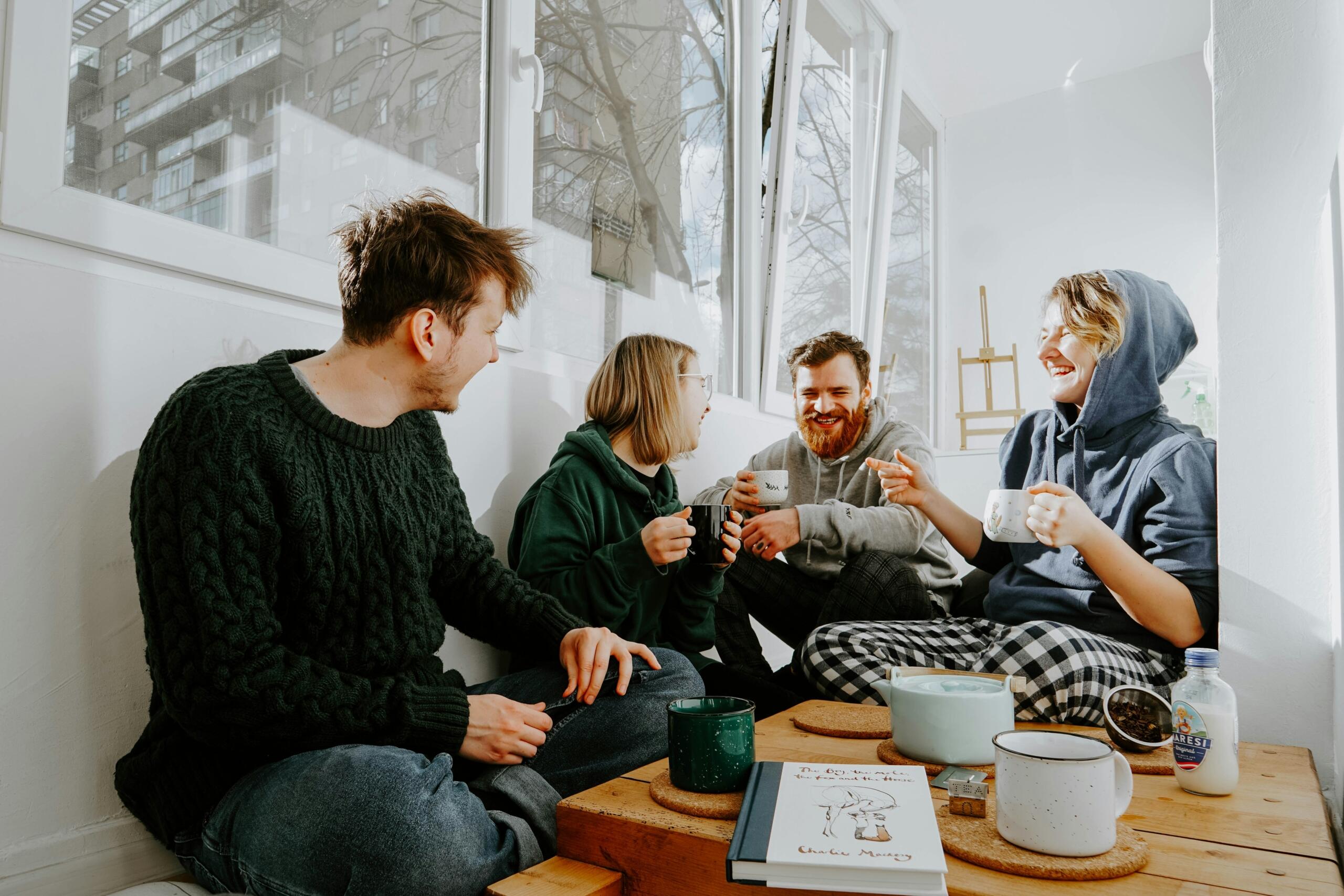 Neli noort täiskasvanut istuvad valgusküllases toas ümber madala laua, joovad kruusidest ning vestlevad omavahel naeratades.