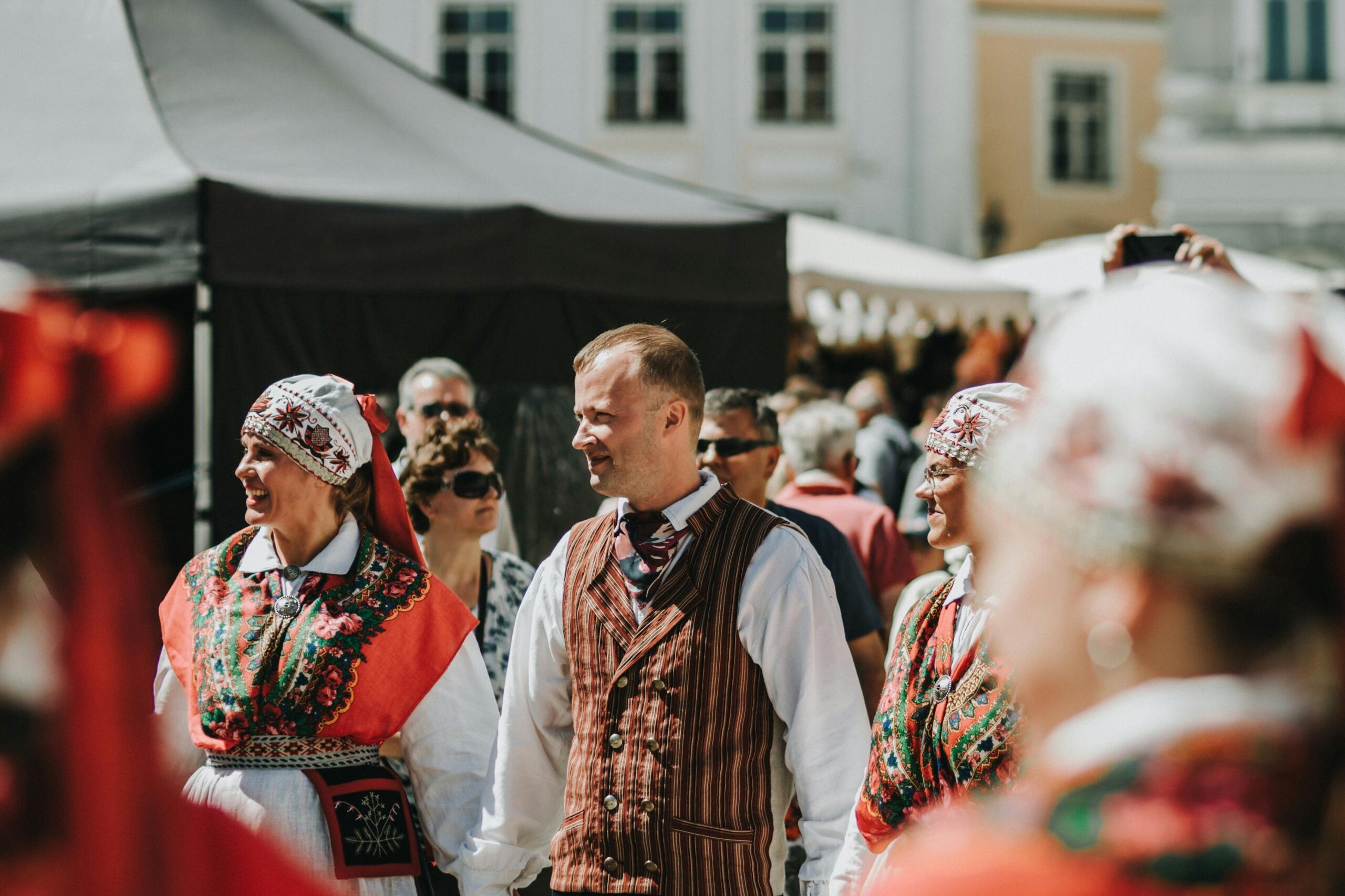 Eesti rahvarõivastes mees ja kaks naist seisavad rahvarohkel väliüritusel.