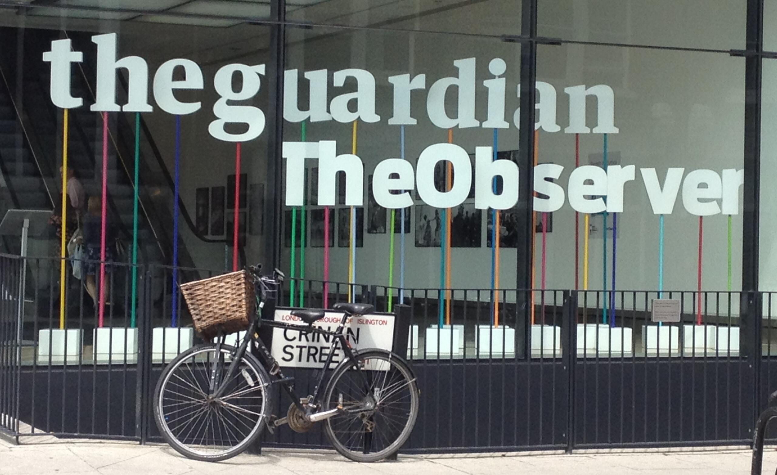 Una bicicleta antigua con una cesta está apoyada contra una barandilla frente a un edificio con grandes ventanales en los que se ven los logotipos de los periódicos The Guardian y The Observer.