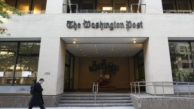 Entrada del edificio de The Washington Post, con una fachada de ladrillo beige, un letrero llamativo y una persona que pasa por allí. El ambiente es profesional y formal.
