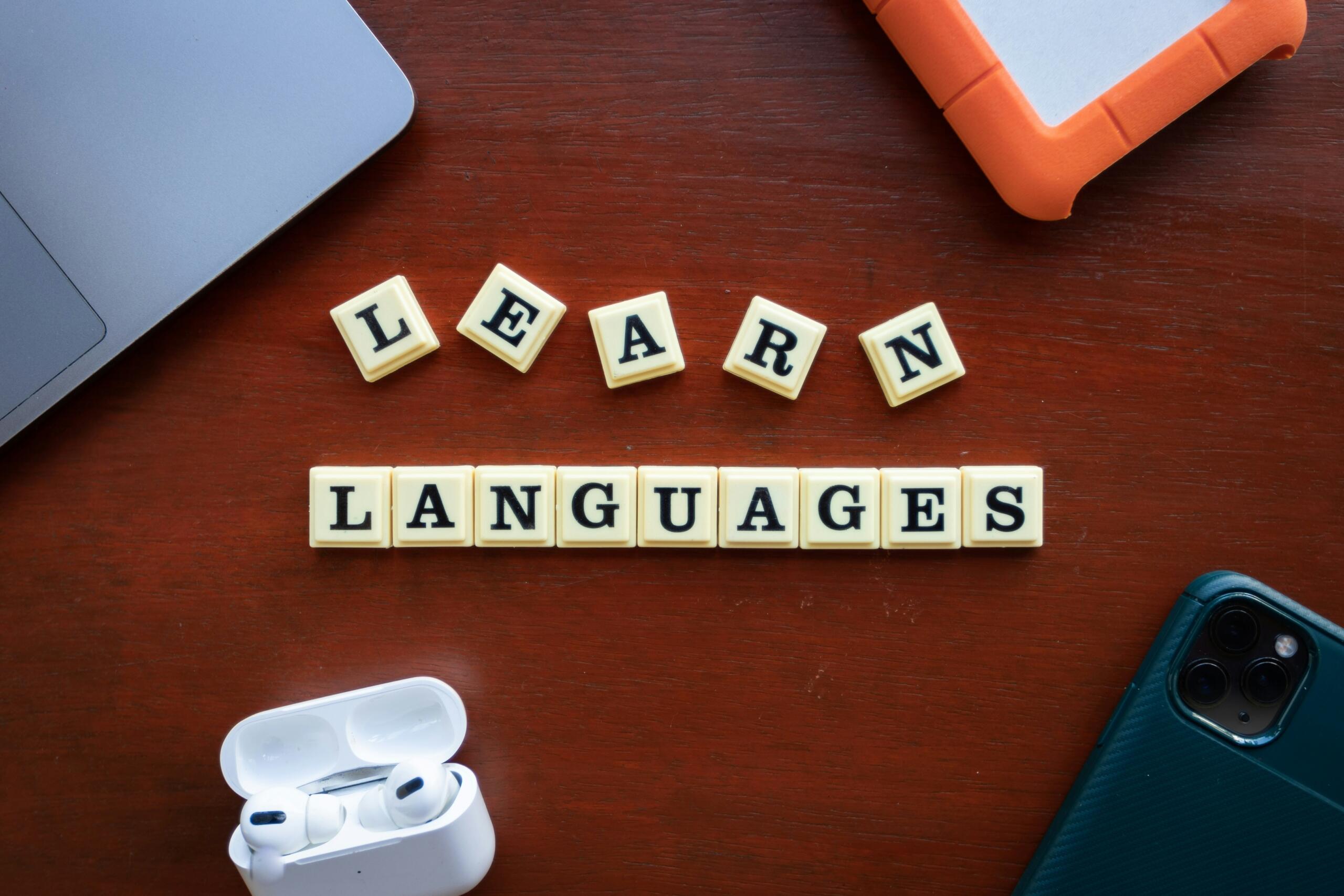 Fichas de Scrabble que forman la frase «LEARN LANGUAGES» sobre un escritorio de madera, rodeadas de un ordenador portátil, un disco duro externo naranja, unos auriculares inalámbricos y un smartphone.