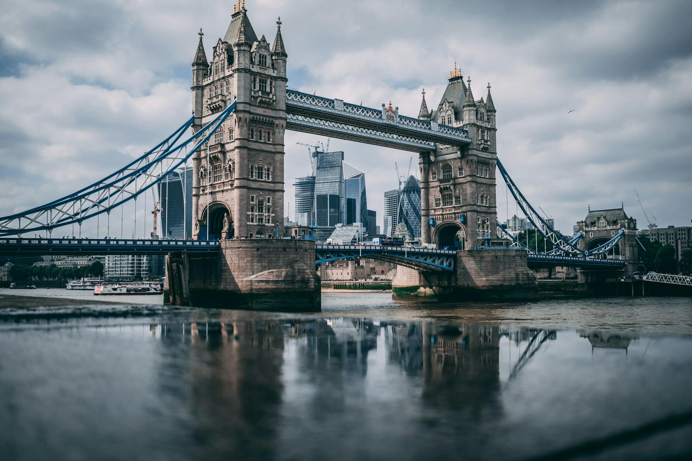 Vista del emblemático puente Tower Bridge de Londres sobre el río Támesis, reflejado en el agua con los rascacielos de la ciudad al fondo bajo un cielo nublado, ideal para representar el inglés británico y sus acentos.