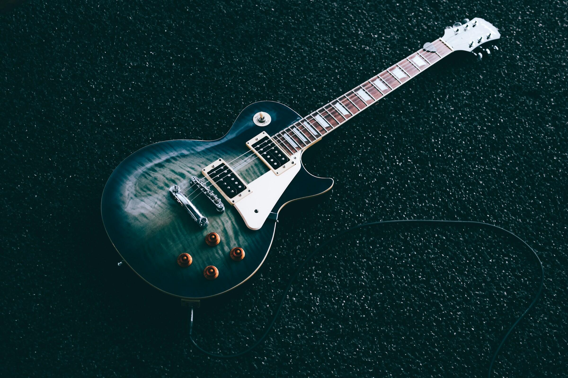 Guitarra eléctrica con acabado azul oscuro y botones marrones sobre una superficie negra texturizada, conectada mediante un cable en espiral.