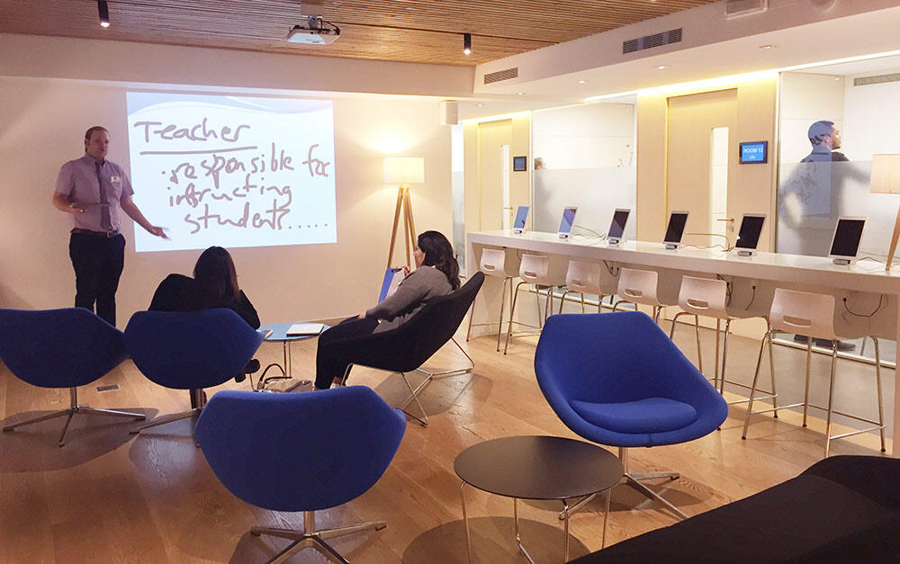 Un profesor se encuentra junto a una pantalla de proyección llena de notas y se dirige a tres alumnos sentados en modernas sillas azules. La sala resulta luminosa y acogedora.