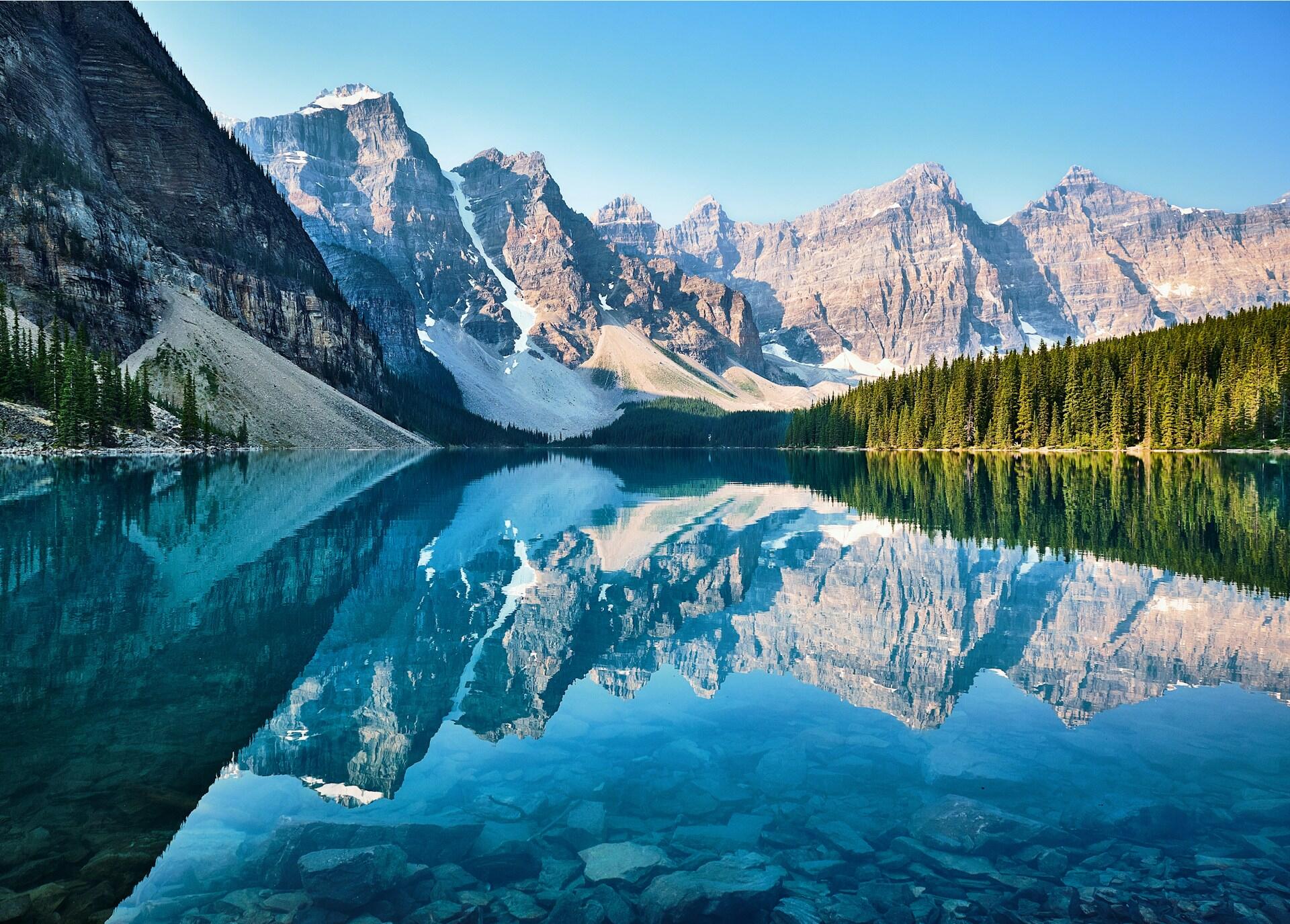 Un lago cristalino en el que se reflejan majestuosas montañas nevadas y un frondoso bosque de coníferas bajo un cielo azul brillante. Un paisaje sereno y tranquilo.