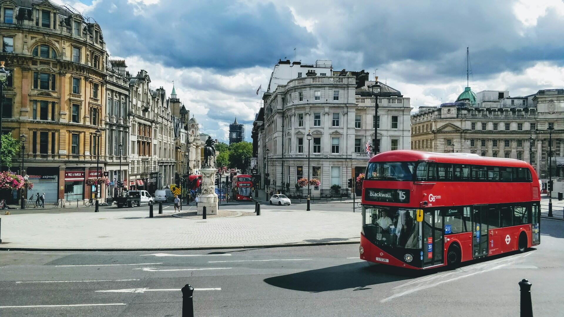 Un autobús rojo de dos pisos circula por una calle ancha en la plaza de una ciudad histórica europea, bajo un cielo nublado y rodeado de edificios antiguos y ornamentados.