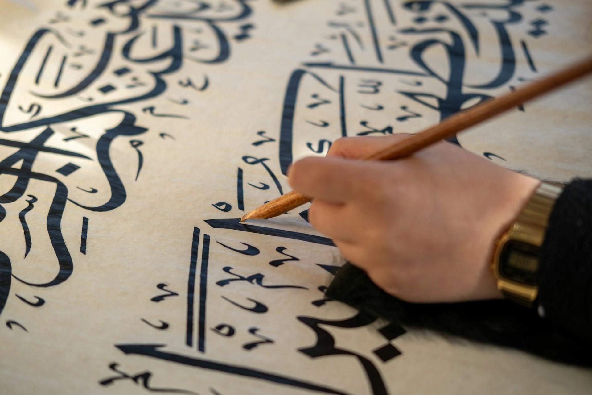 Mano practicando caligrafía árabe con un lápiz sobre papel.