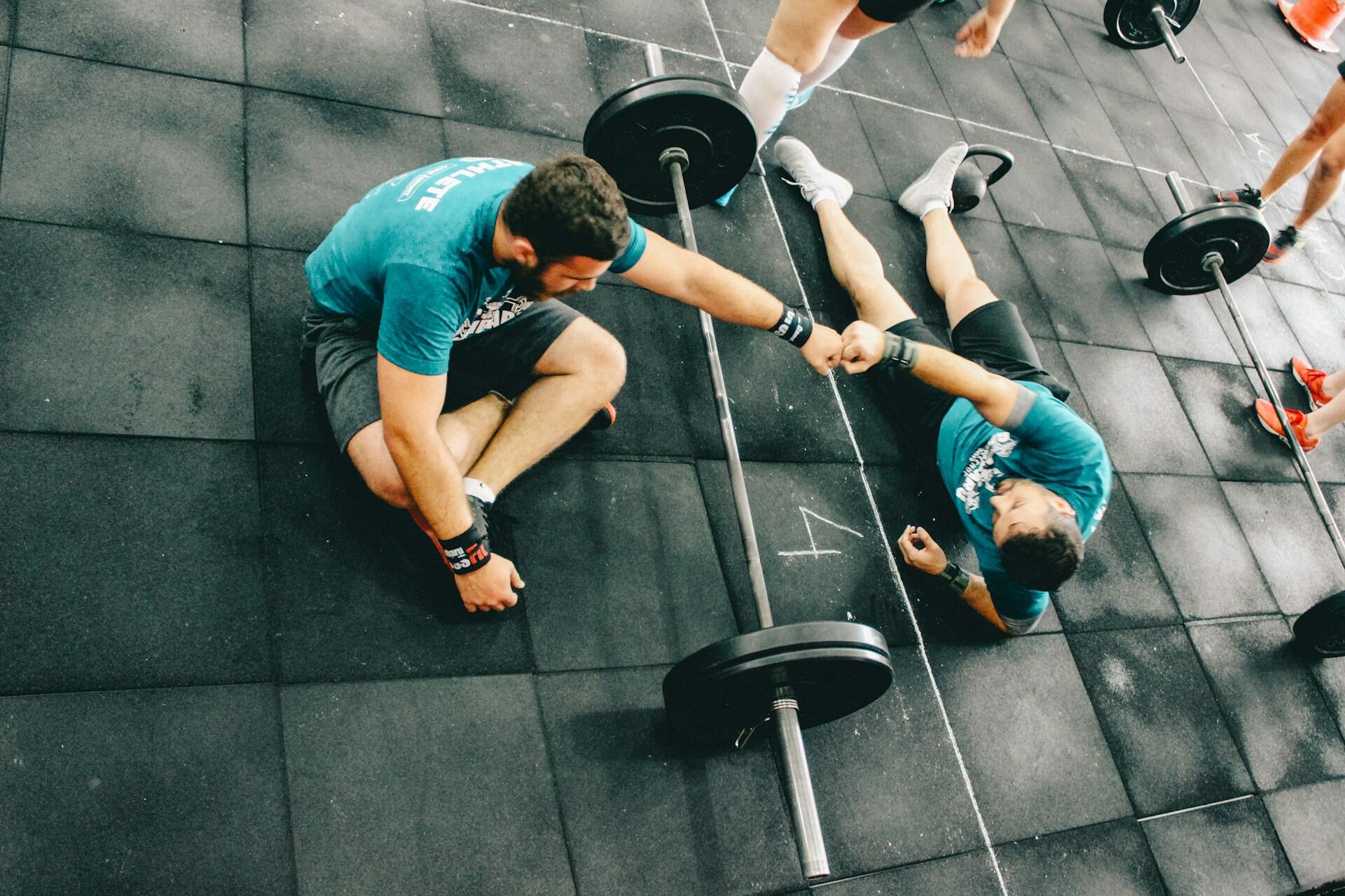 Dos hombres con camisetas verde azulado en un gimnasio están sentados en el suelo junto a una barra, dándose la mano en señal de camaradería. La escena transmite trabajo en equipo y apoyo.