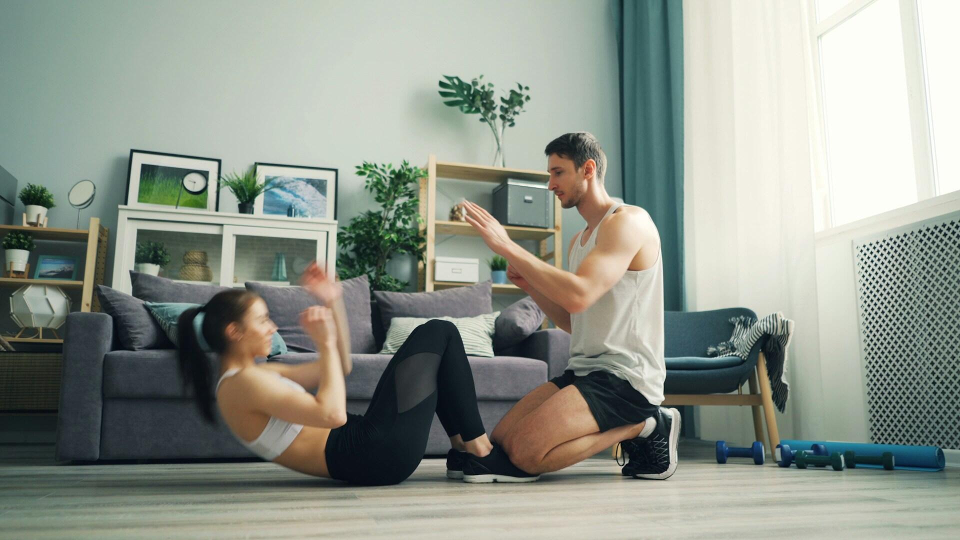 Un hombre ayuda a una mujer mientras hace ejercicio en casa; ella está haciendo abdominales sobre un suelo de madera. La habitación es luminosa y moderna, con un sofá y estanterías.
