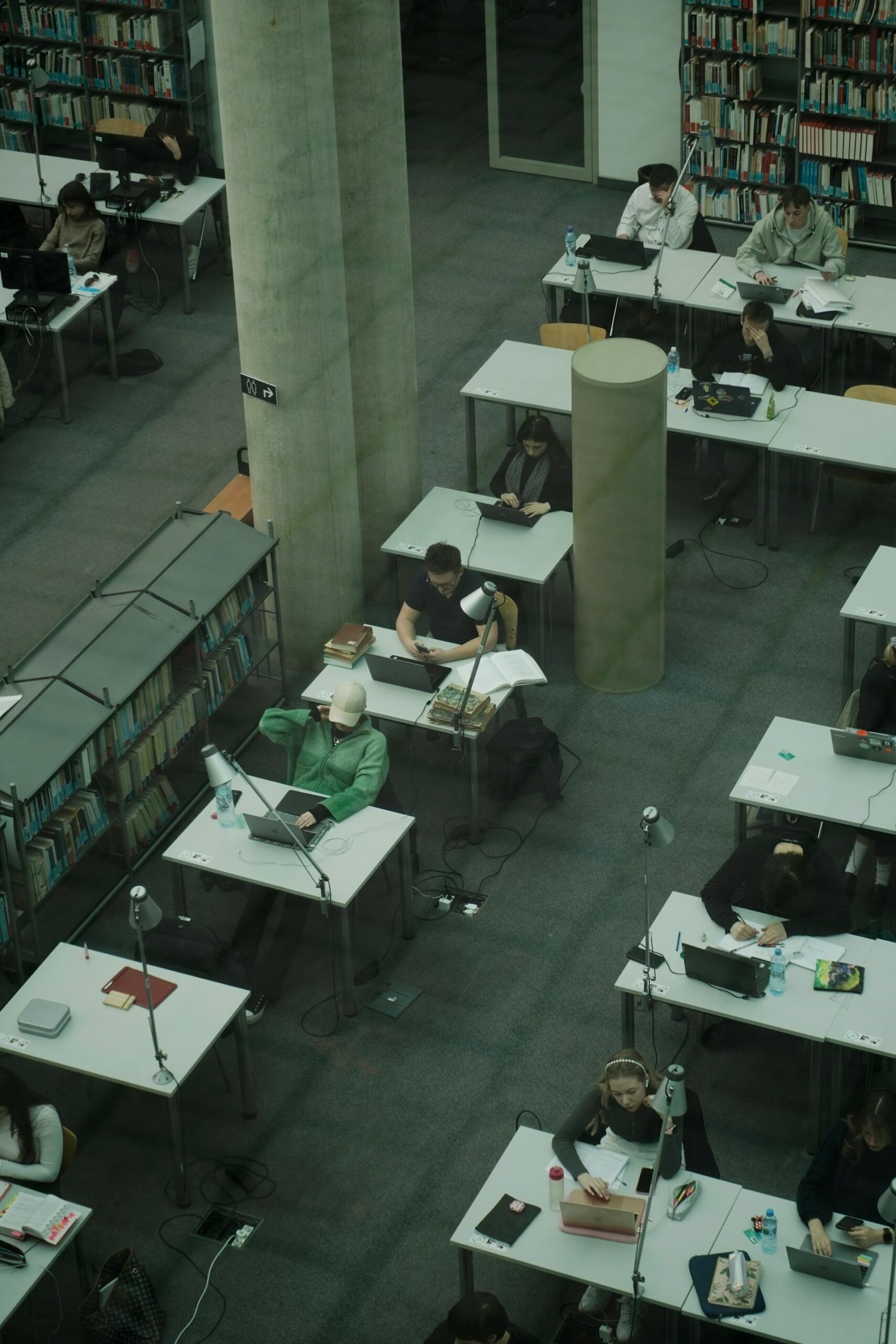 Vista aérea de una biblioteca en la que se ve a gente estudiando en las mesas, utilizando ordenadores portátiles y leyendo libros. El ambiente es tranquilo y propicio para la concentración, y las paredes están repletas de estanterías.