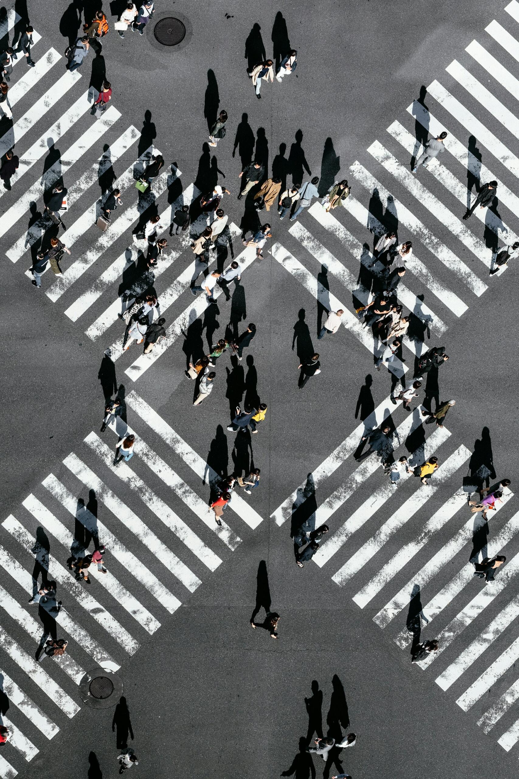 Vista aérea de un cruce muy transitado con pasos de cebra en diagonal, repletos de peatones que proyectan largas sombras en un día soleado.