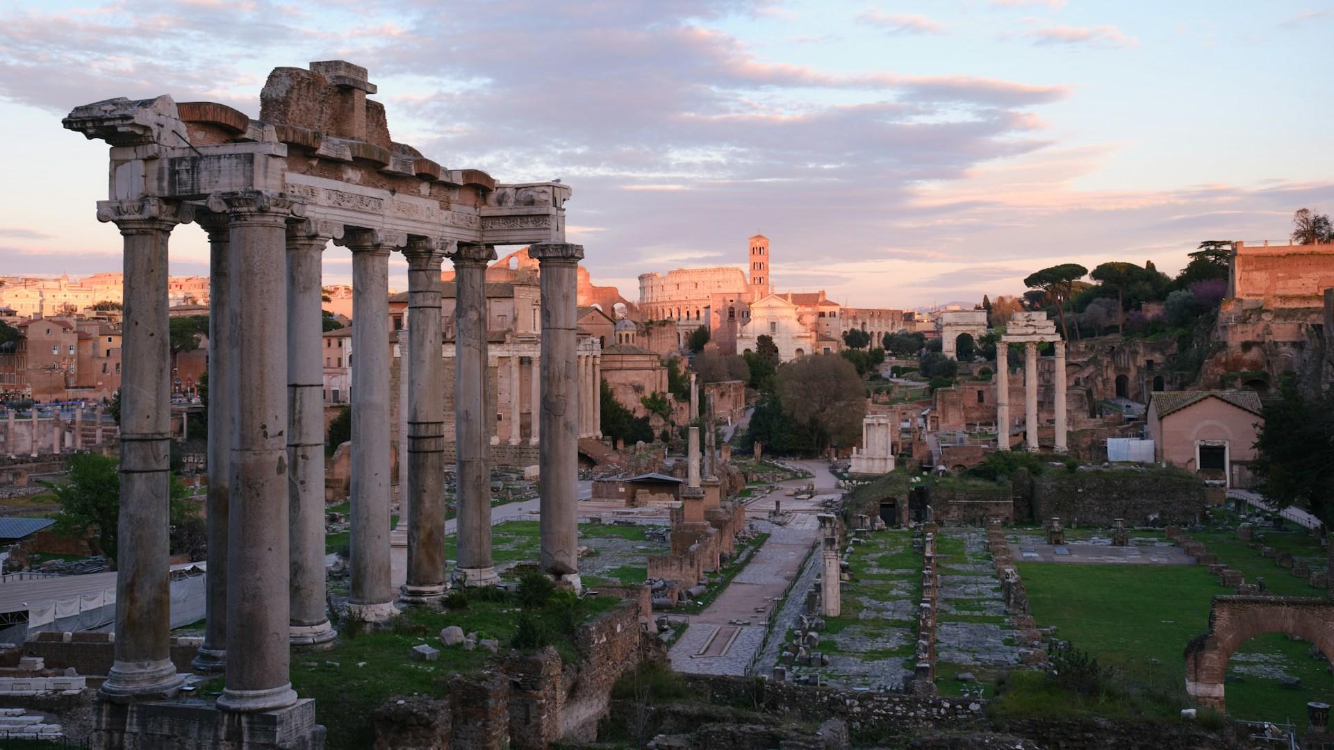 Antiguas ruinas romanas bañadas por los tonos del atardecer con el Coliseo al fondo.