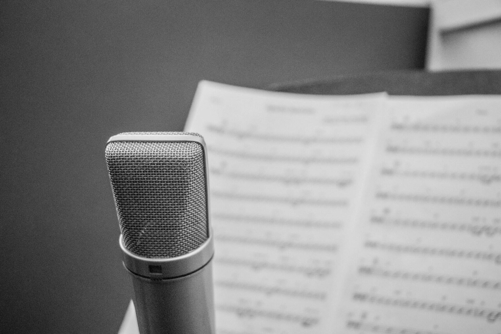 Primer plano de un micrófono delante de una partitura desenfocada. La imagen en blanco y negro transmite una sensación de concentración y creatividad en un estudio de grabación.