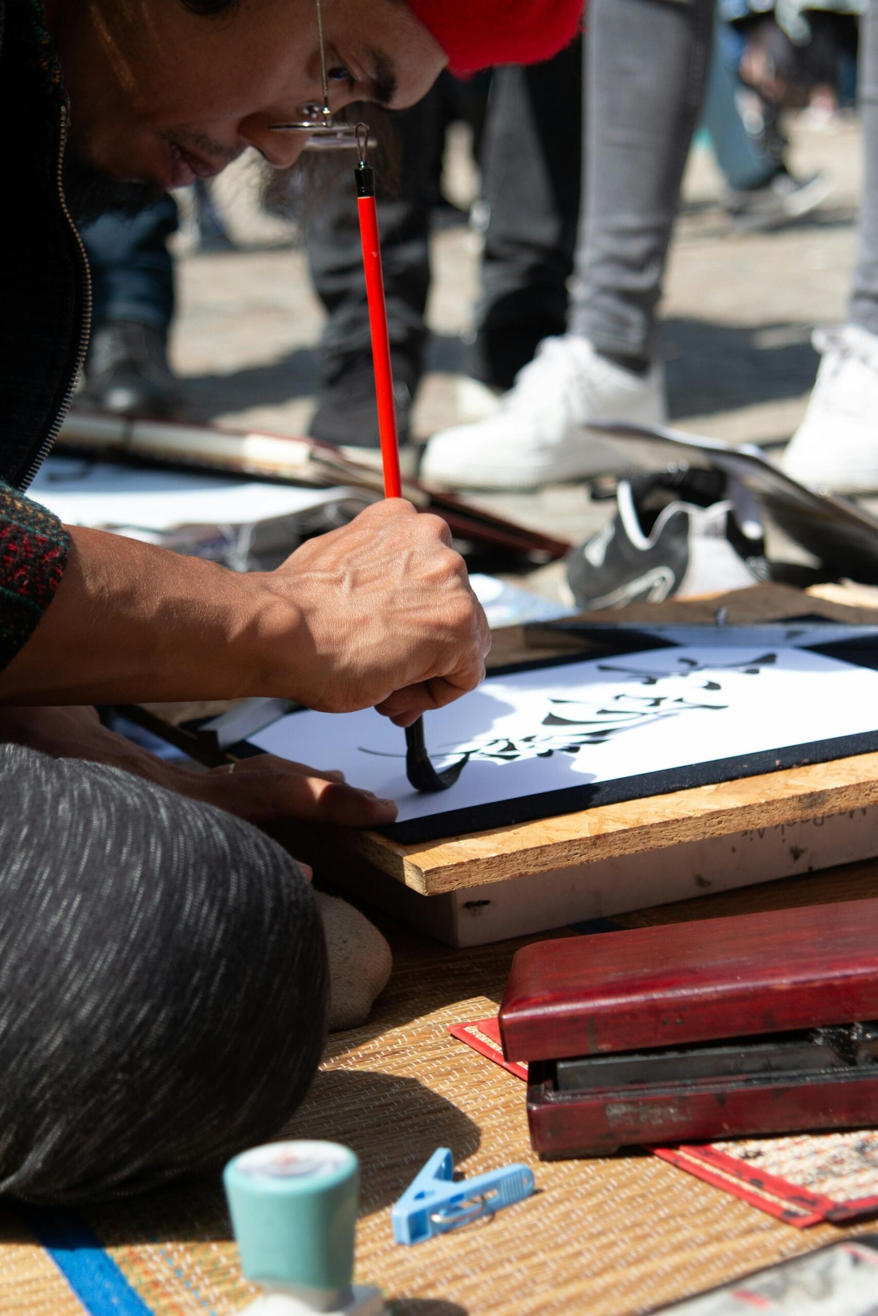Un artista pinta a mano letras caligráficas negras sobre un cartel blanco al aire libre, rodeado de herramientas y materiales de pintura; ambiente de taller callejero, creativo y de gran concentración.